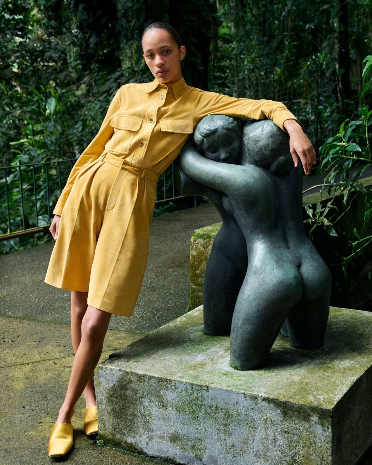 Woman in yellow outfit posing next to a modern sculpture of two intertwined figures in a lush, green outdoor setting.