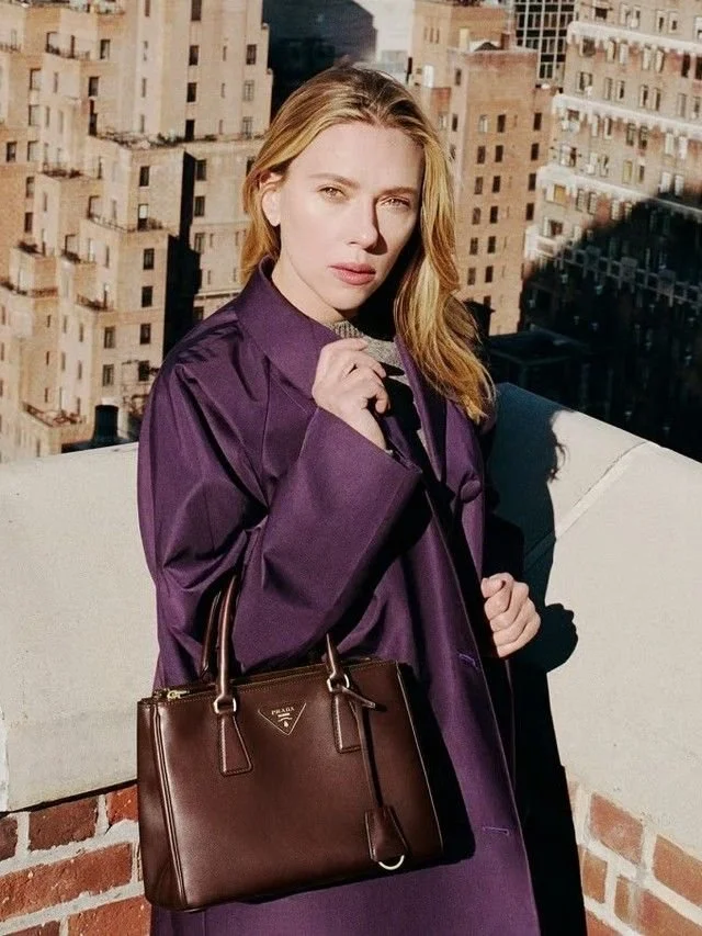 A woman with blond hair stands outdoors on a rooftop with a city skyline in the background. She wears a purple coat and carries a brown Prada handbag.