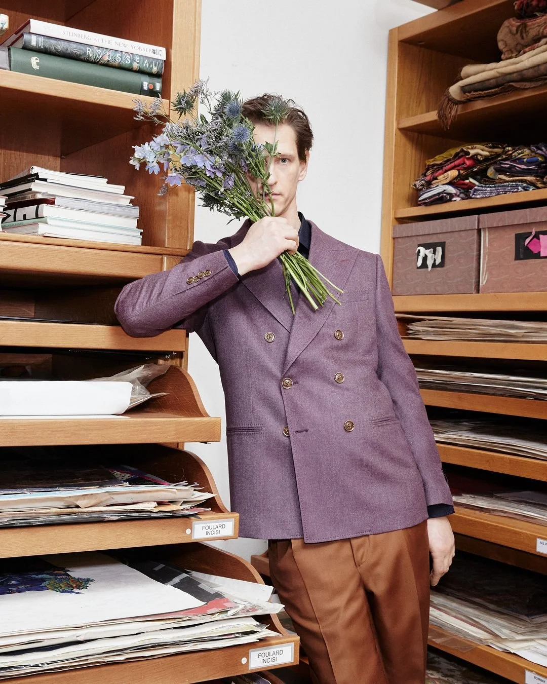 A man in a purple double-breasted blazer holding a bouquet of purple and blue flowers in front of his face, standing in a room with shelves filled with books, papers, and fabric.