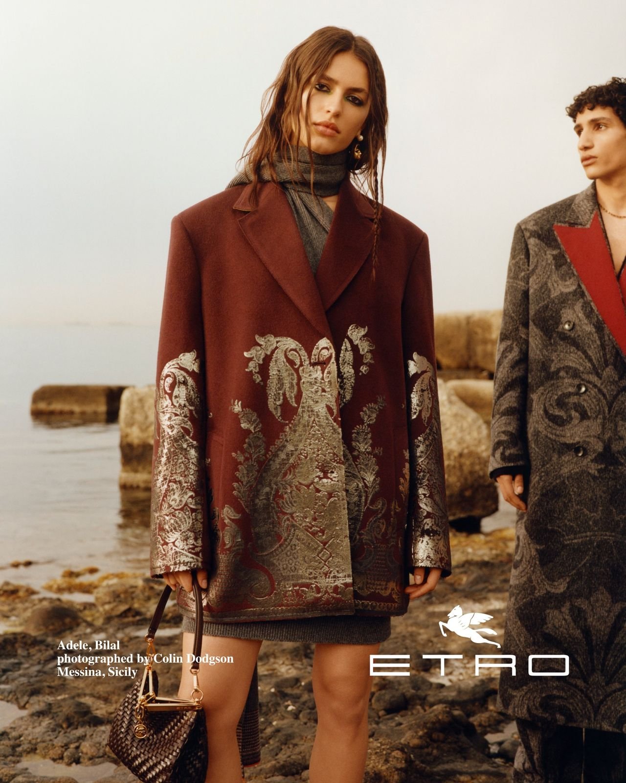 Fashionable woman standing on rocks by the water, wearing a patterned coat with elaborate embroidery, holding a small handbag, with another person partially visible beside her. The scene is set at a beach or seaside.