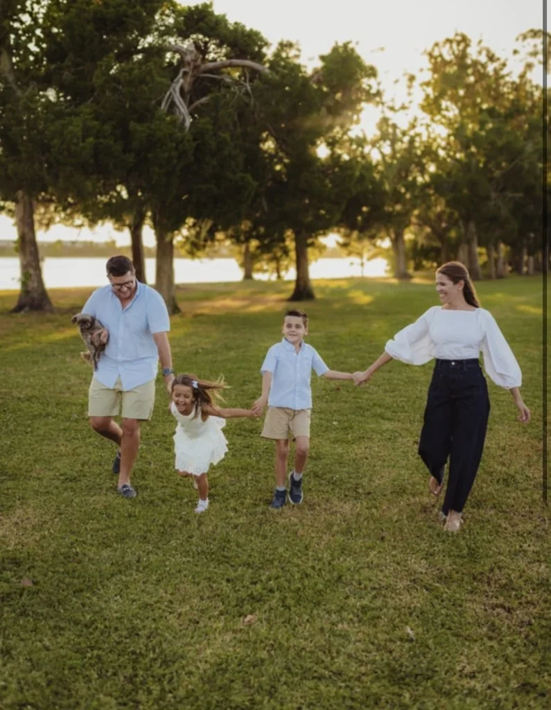 A family of four, including two children, is running on a grassy field holding hands, with trees and a river in the background during sunset.