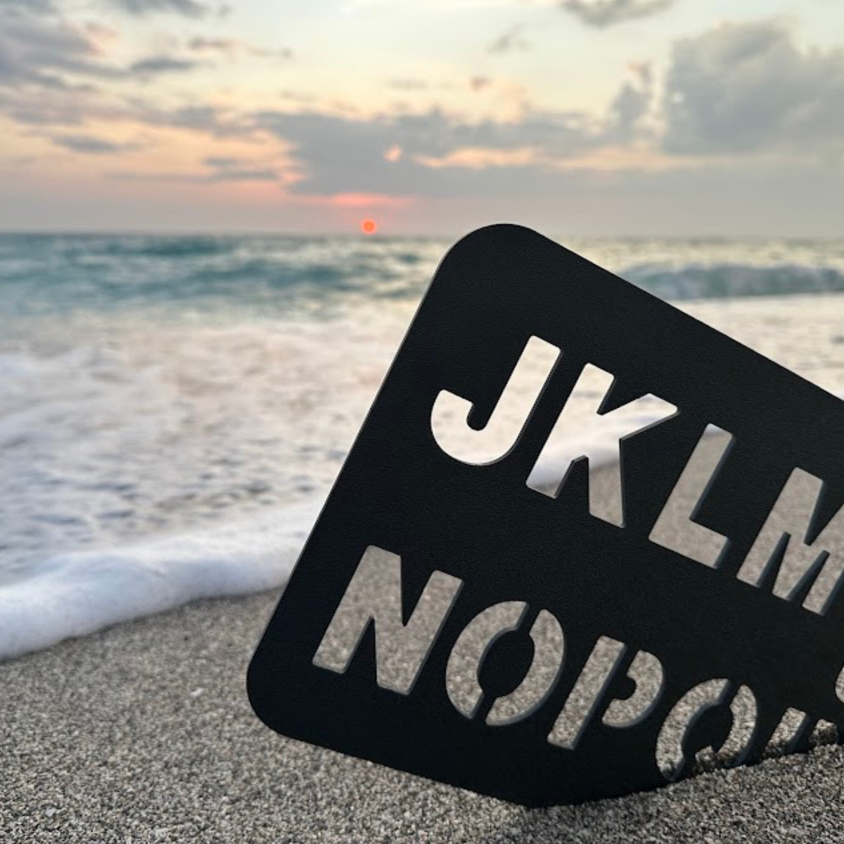 A sand beach with ocean waves and a setting sun in the background, with a black sign that reads 'JACKSON' resting on the sand.