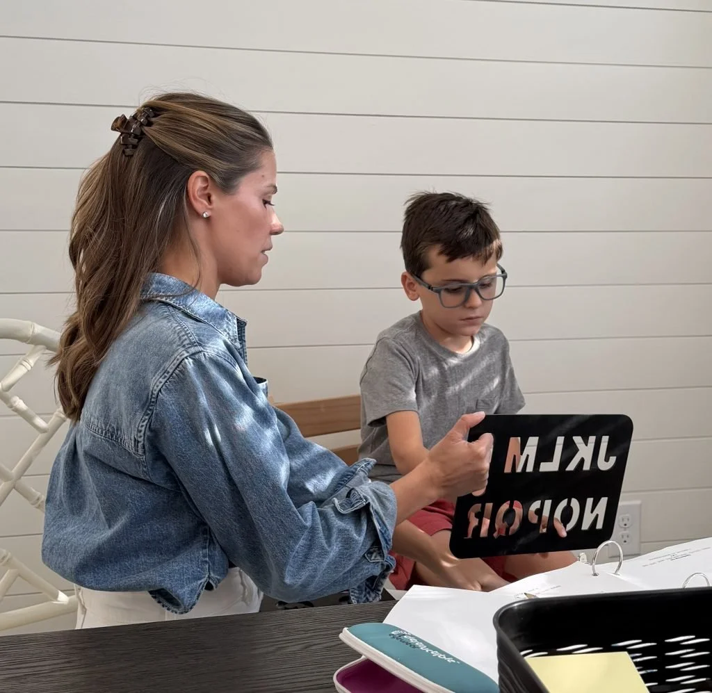A woman sitting at a table holding a chalkboard sign that says 'MKLM' and 'WORLD' in mirror image, while a boy with glasses sits beside her looking at it, in a room with white wooden-paneled walls.