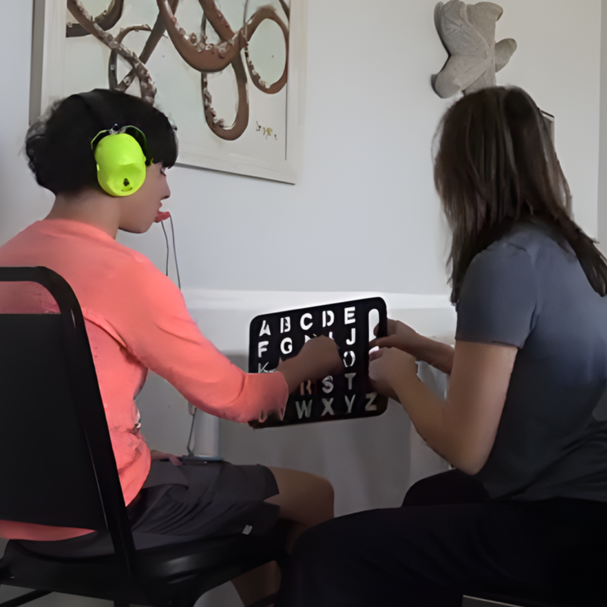 A young girl with headphones and a woman sit at a table, playing a word or letter game with a black board displaying the alphabet. The girl wears a pink top and gray shorts, and the woman wears a gray shirt. There is abstract art on the wall behind them.