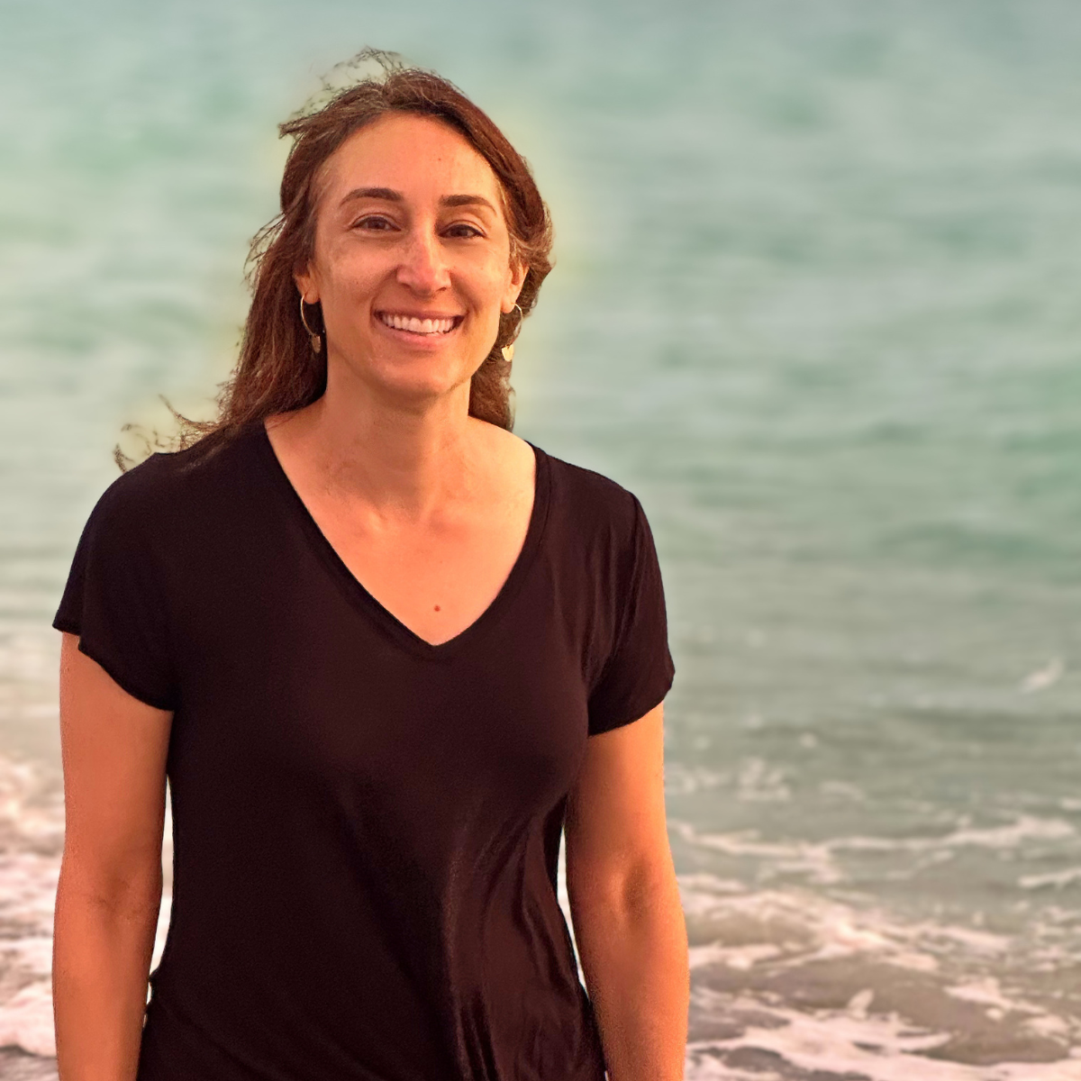 A woman with brown hair smiling at the camera, standing on a beach with the ocean in the background.