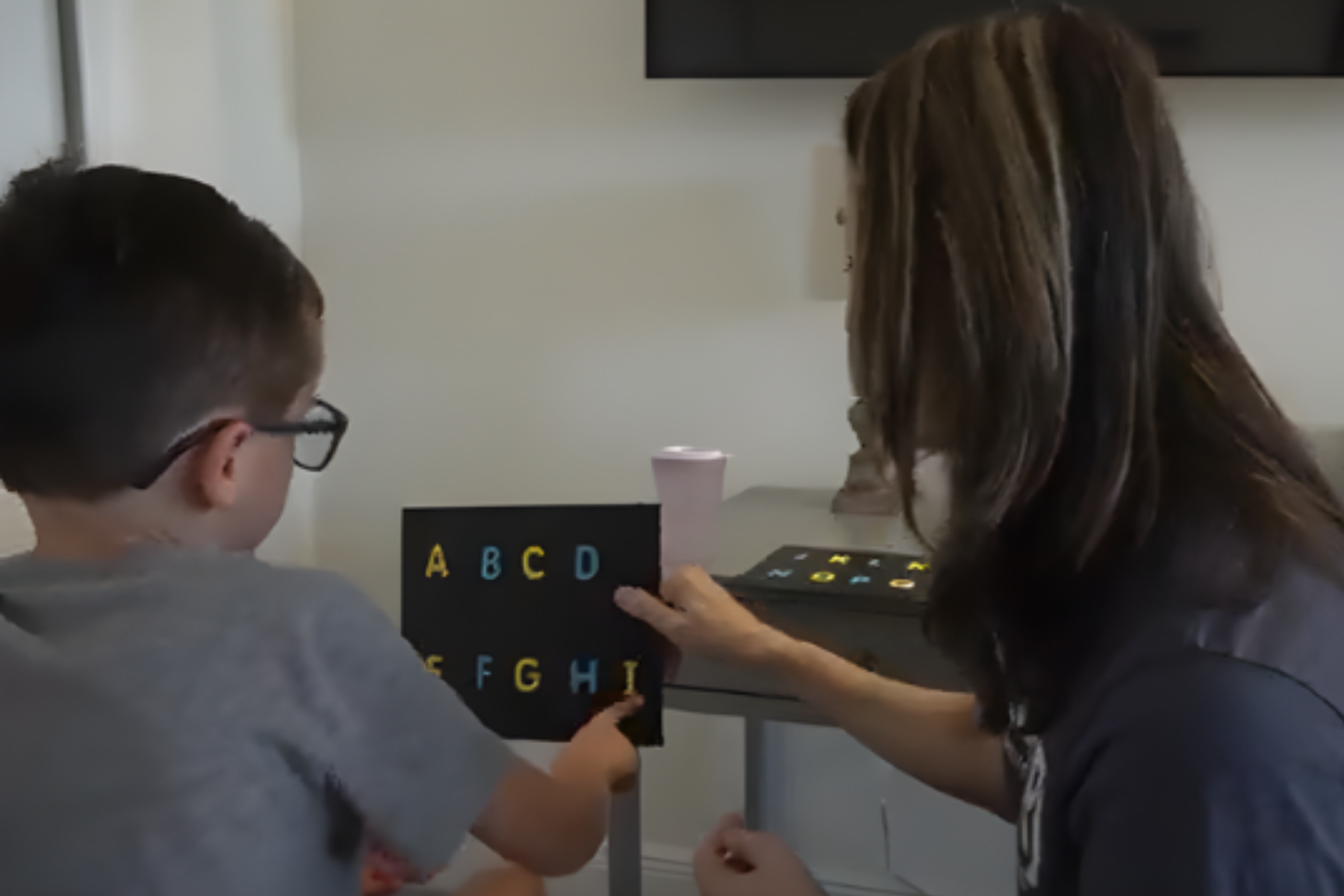 A young boy with glasses and an adult woman sitting at a table, holding a black letterboard with colorful letters 'A B C D' on it, in a room with a TV and a pink cup in the background.