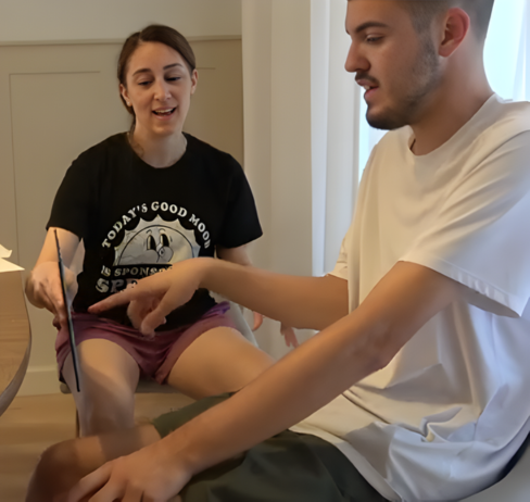 Two young adults sitting indoors, one smiling and pointing at the other's arm, which the other person is touching.