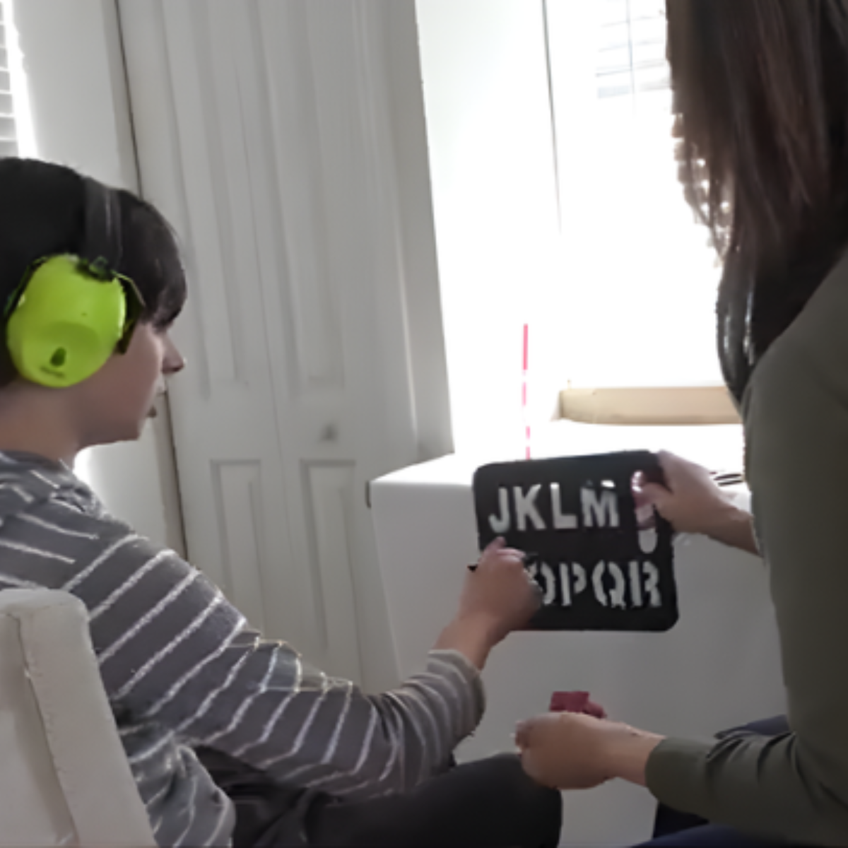 A child wearing green earmuffs is sitting in a chair and looking at a device, while an adult holds a letter board with the alphabet.