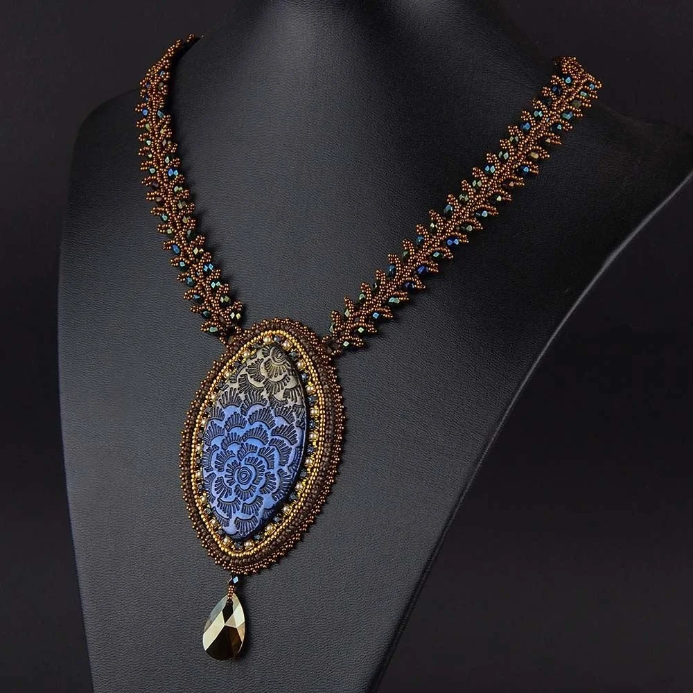 Elegant necklace with a large oval black and gold patterned pendant, surrounded by gold and blue beads, and a teardrop-shaped black gemstone hanging at the bottom, displayed on a black bust.