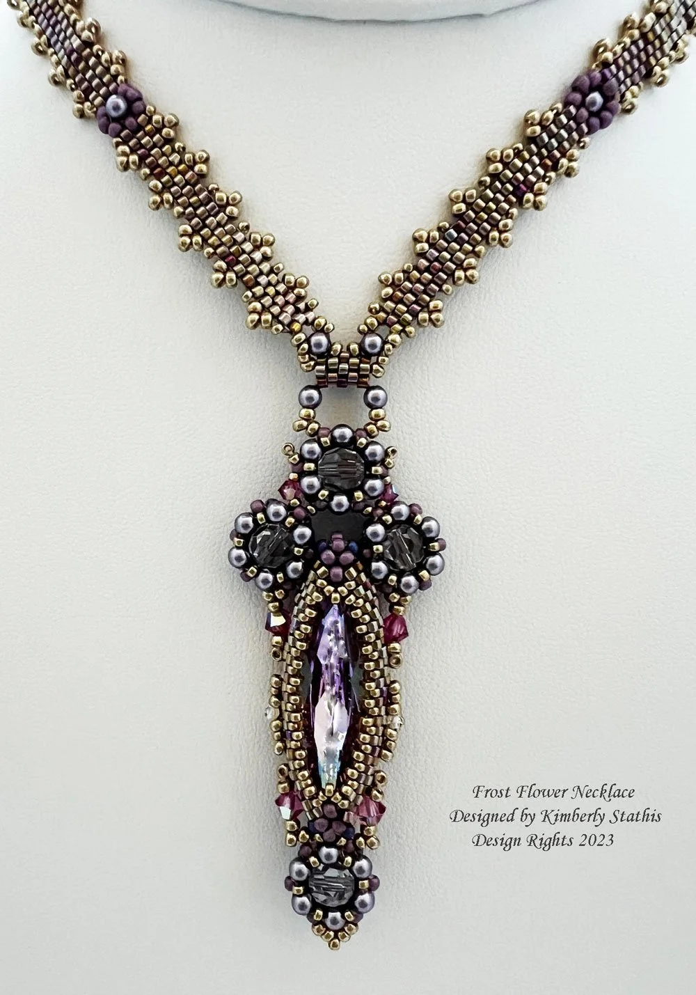 Beaded necklace named Frost Flower, designed by Kimberly Stathis, with purple, black, gold, and pearl beads, and a large central purple gemstone, hanging against a white background.