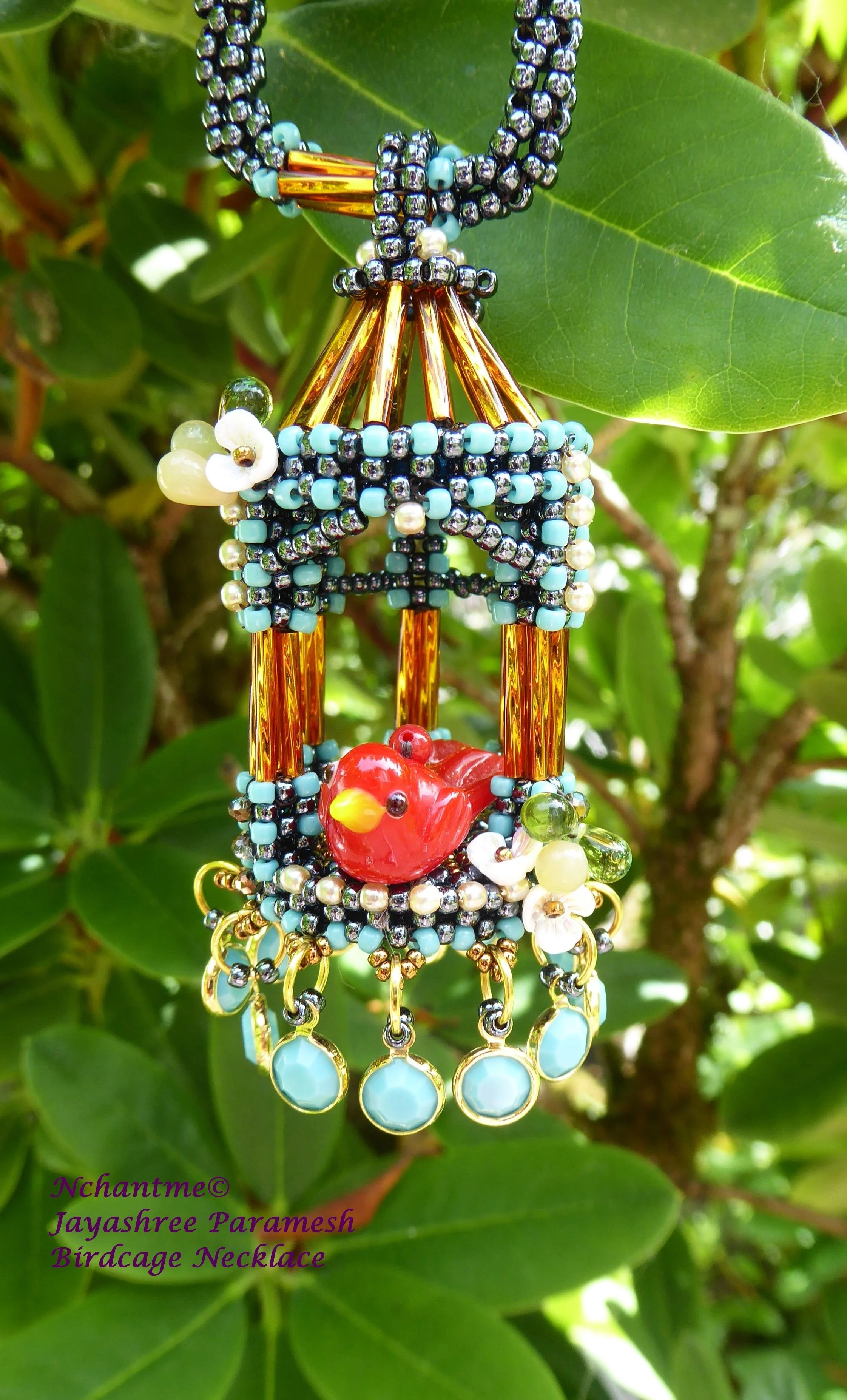 Close-up of a beaded birdcage-shaped necklace hanging on a green leafy plant. The necklace features small beads in black, white, blue, and gold, with a red bird pendant at the center. Text in purple at the bottom left corner reads: 'Nchantme© Jayashr