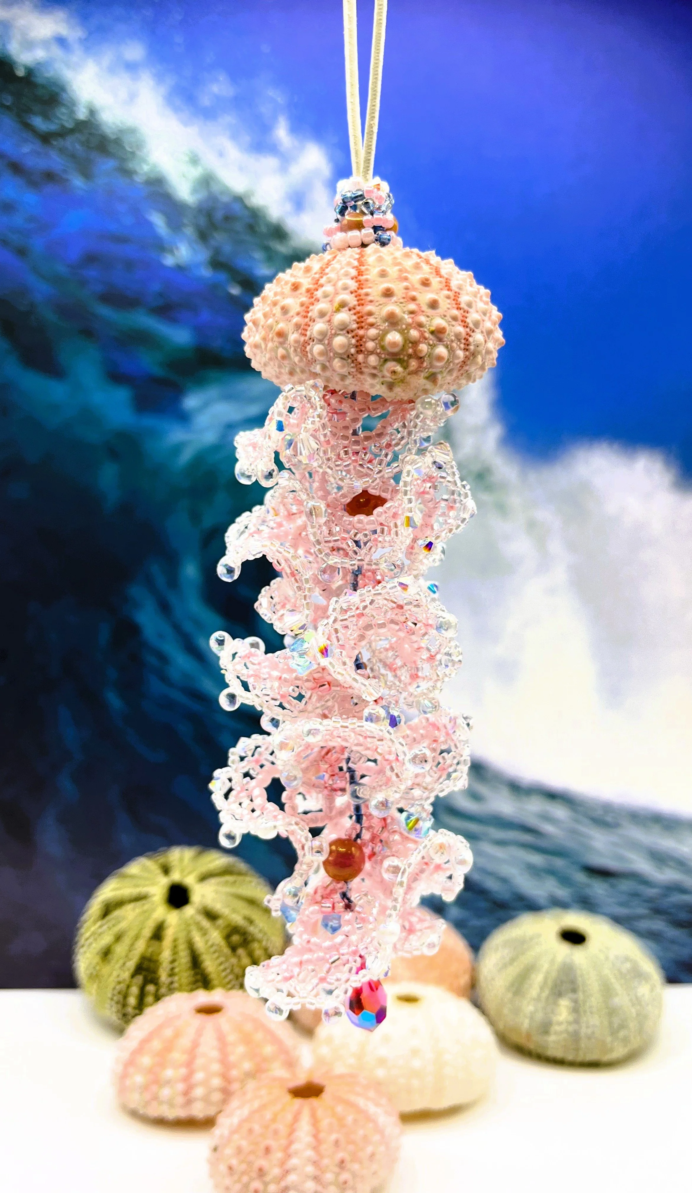 A beaded jewelry piece hanging from a string with seashells and coral in the background, set against a blue backdrop that resembles ocean waves.