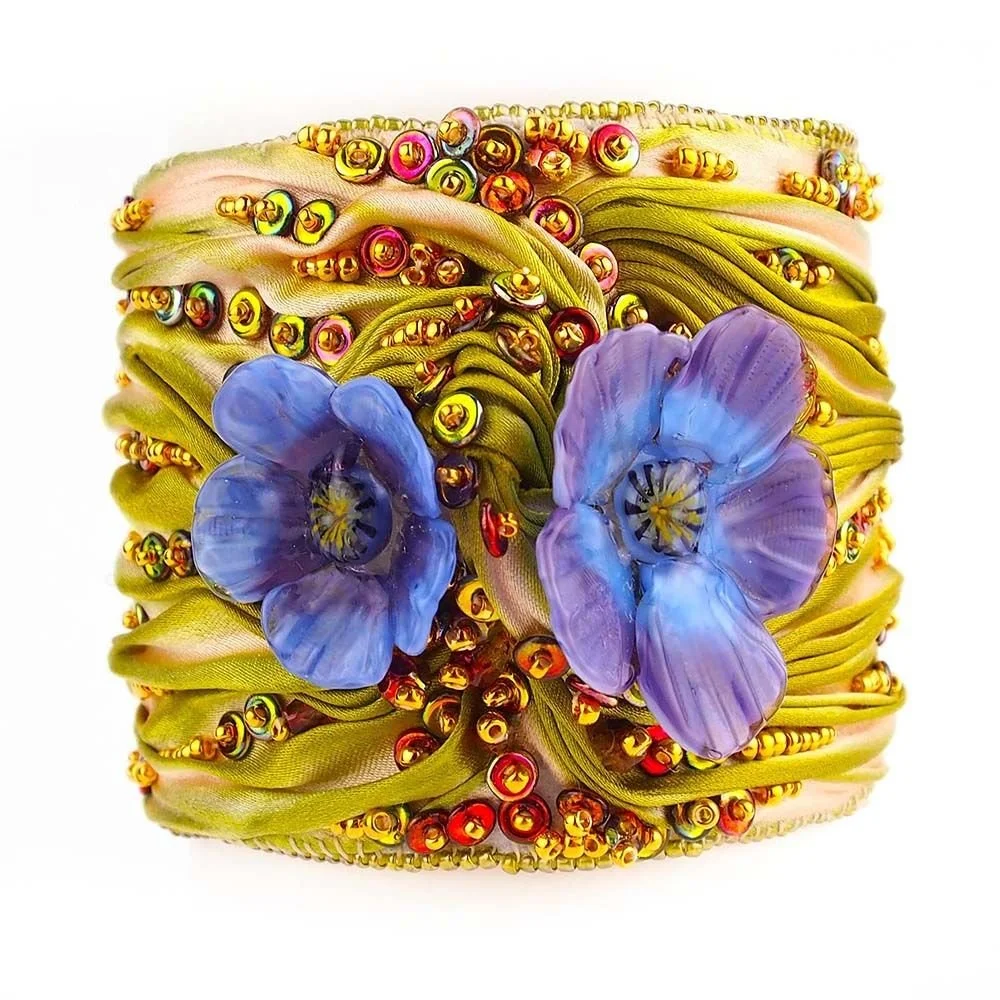 A decorative bracelet with purple flowers, gold beads, and green fabric ribbons.
