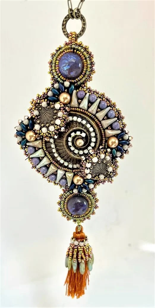 Colorful, intricate handcrafted pendant with beads, pearls, and a spiral design, hanging from a chain with orange tassel at the bottom.
