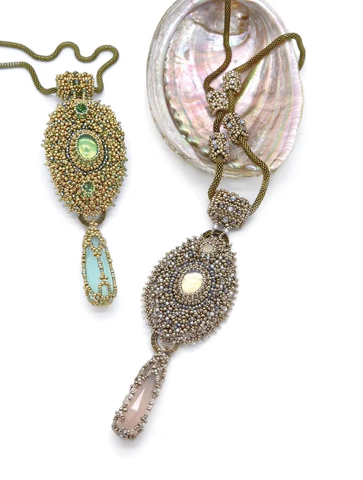 Two ornate necklaces with large oval gemstones and intricate beadwork, one in gold and pastel colors, the other in silver and pastel colors, displayed on a white surface near an open shell.