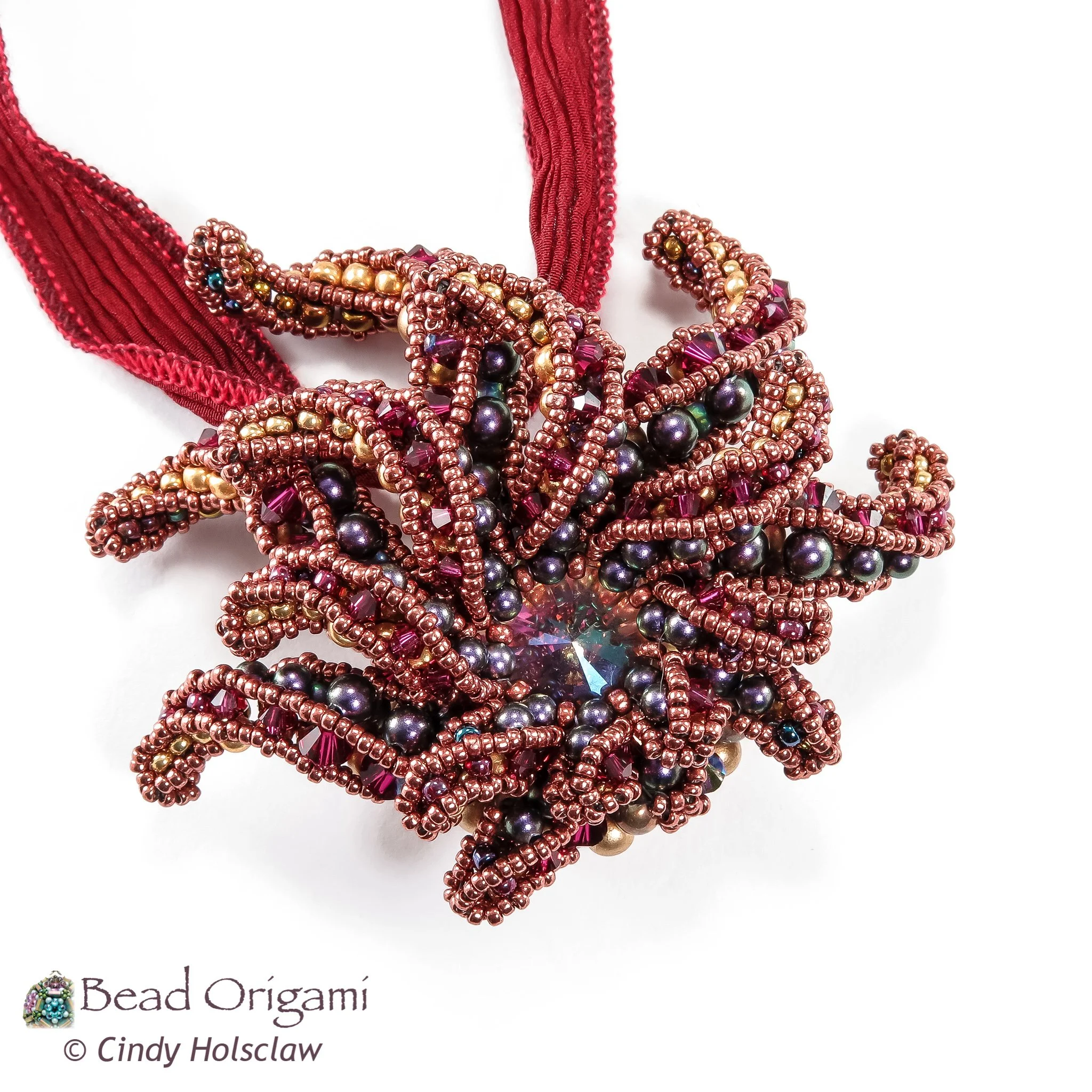Close-up of a beaded jewelry piece with pink, purple, gold, and black beads, attached to a pink fabric ribbon, on a white background.