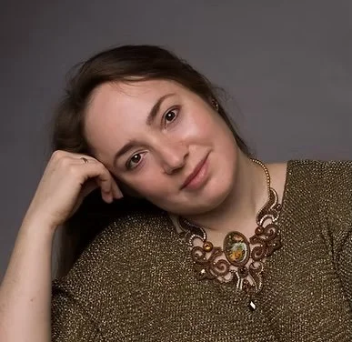 Portrait of a woman with brown hair, resting her head on her hand, wearing a gold top and an ornate statement necklace.