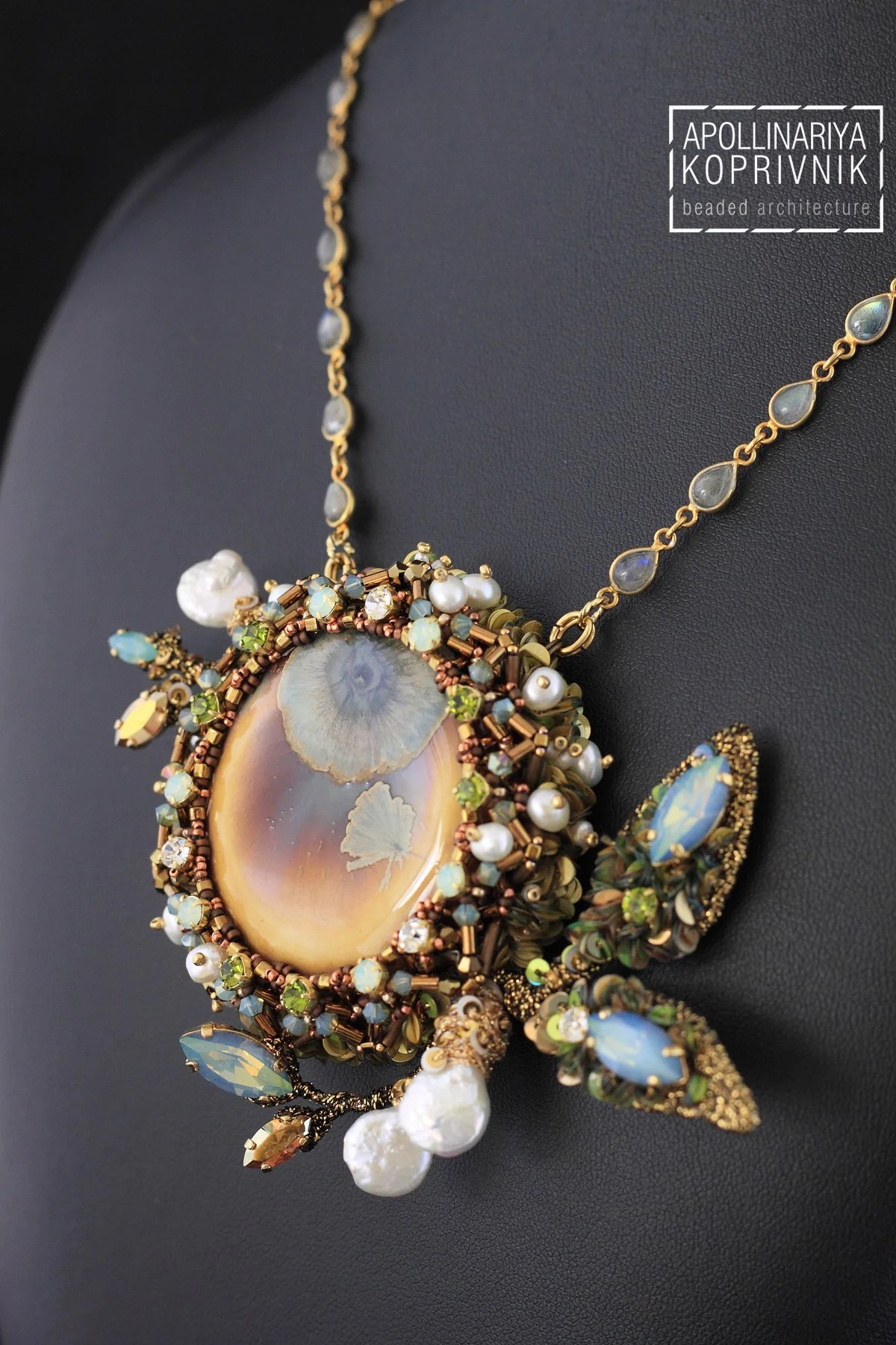 Close-up of an ornate pendant necklace featuring a large opal stone surrounded by small beads, pearls, and gemstones with bee and butterfly motifs, resting on a black display stand.