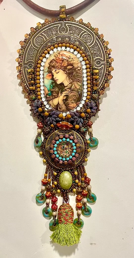 Beaded decorative necklace with a pendant featuring an illustration of a woman with long flowing hair, surrounded by various beads, stones, and charms, ending with a yellow tassel.