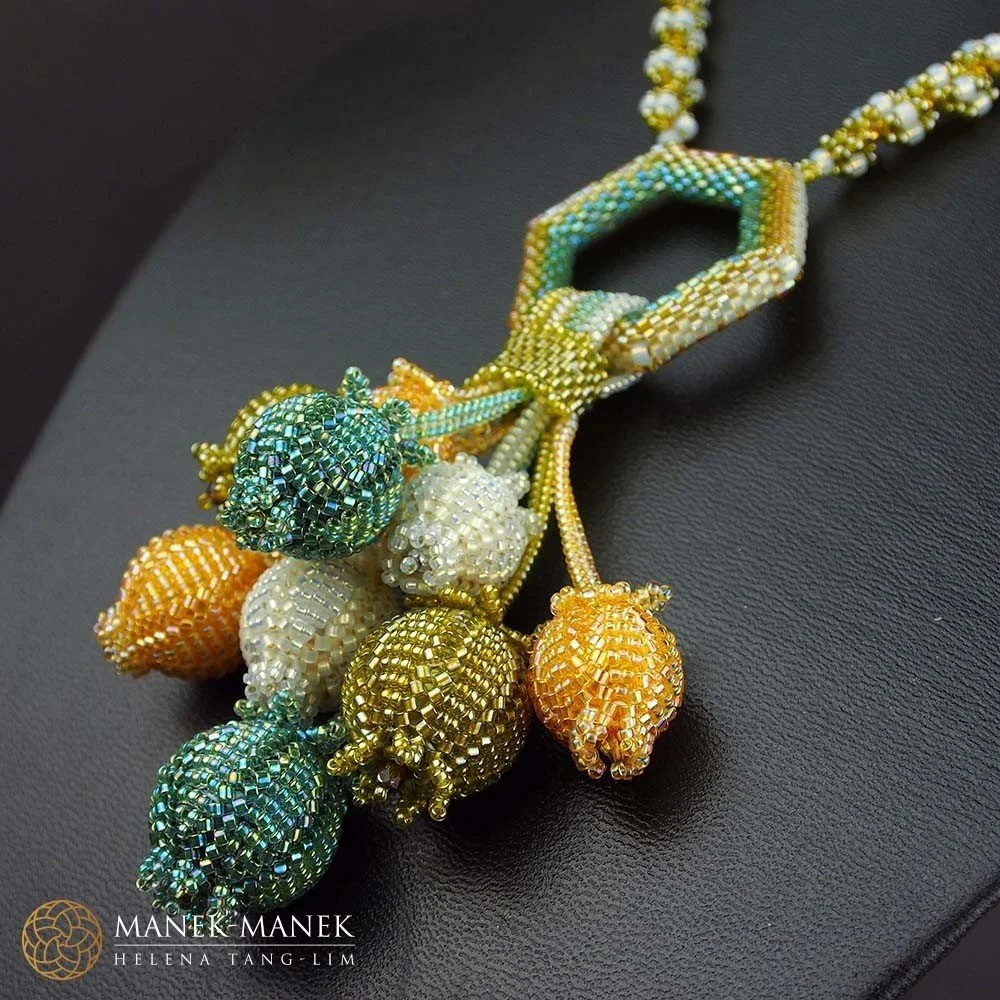 Colorful beaded necklace featuring a geometric pendant and fruit-shaped beads in green, orange, and white, resting on a black surface.