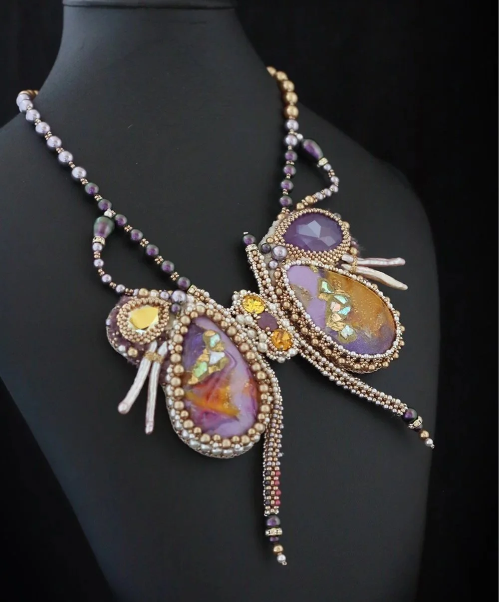A necklace with large purple, yellow, and iridescent stones, surrounded by small pearls and beads, displayed on a black mannequin.