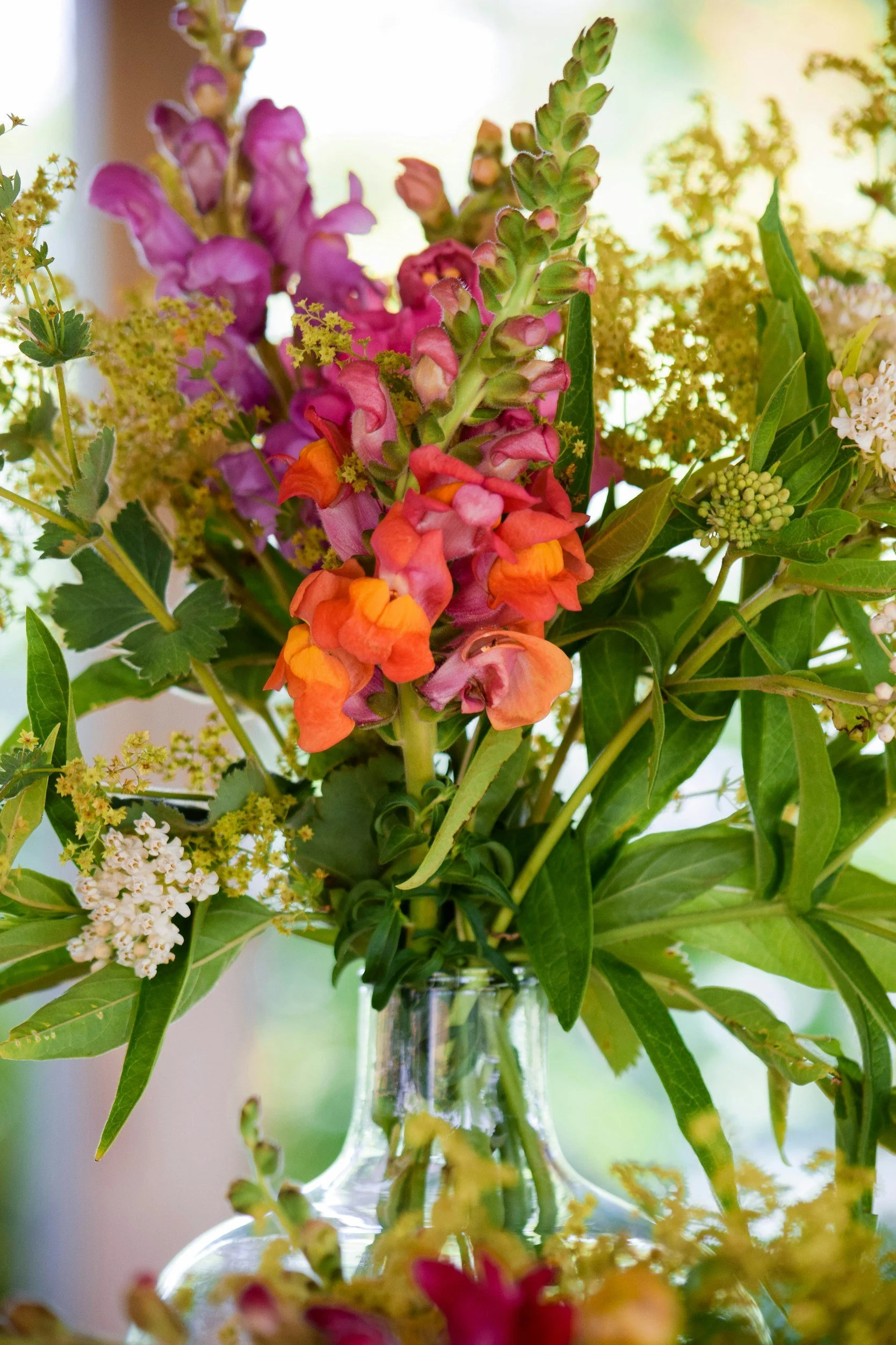 A close-up of a colorful flower bouquet in a vase, featuring pink, purple, orange, and yellow flowers with green leaves.