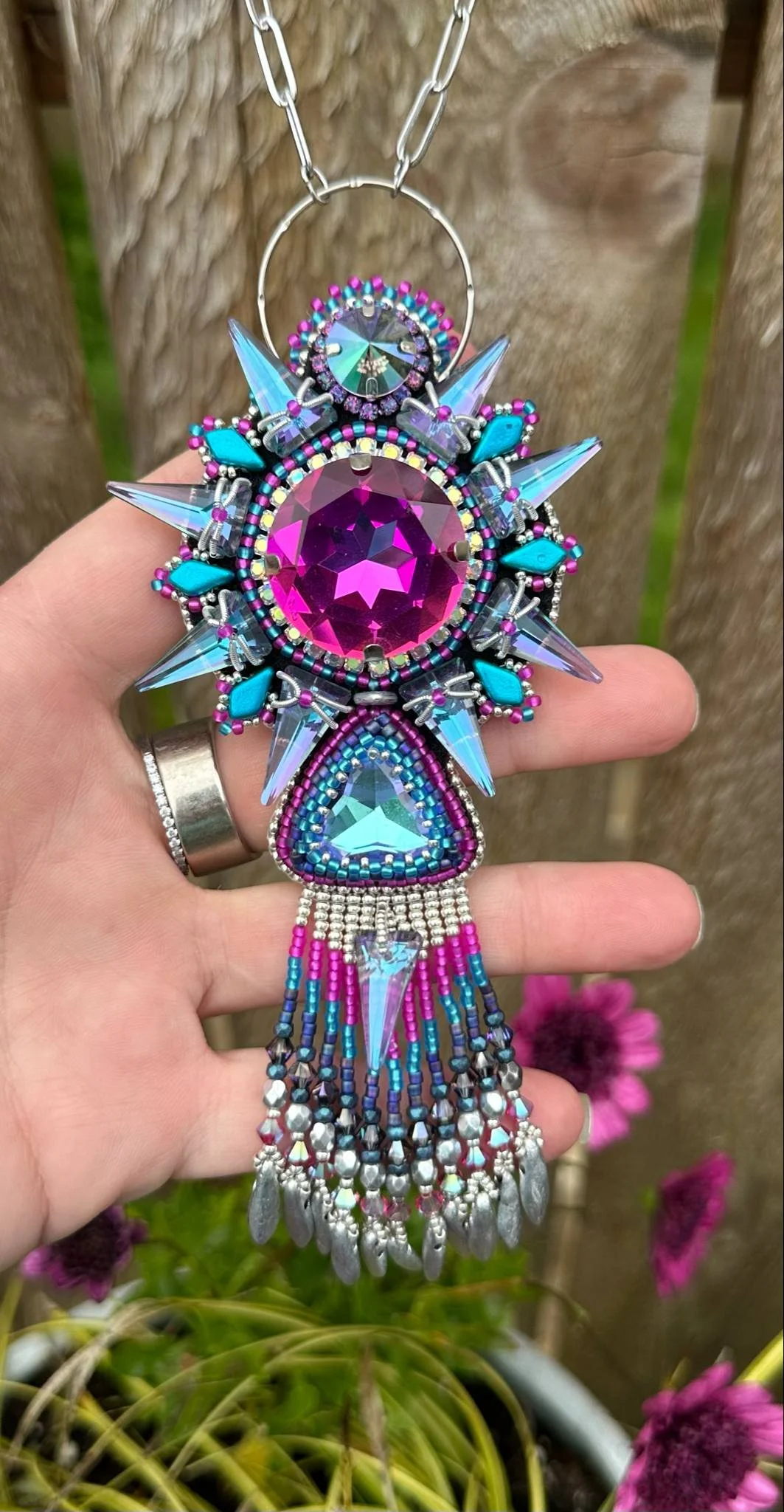 Hand holding a large, colorful beaded and jeweled necklace with pink, blue, purple, and silver accents, hanging in front of a wooden background with pink flowers.