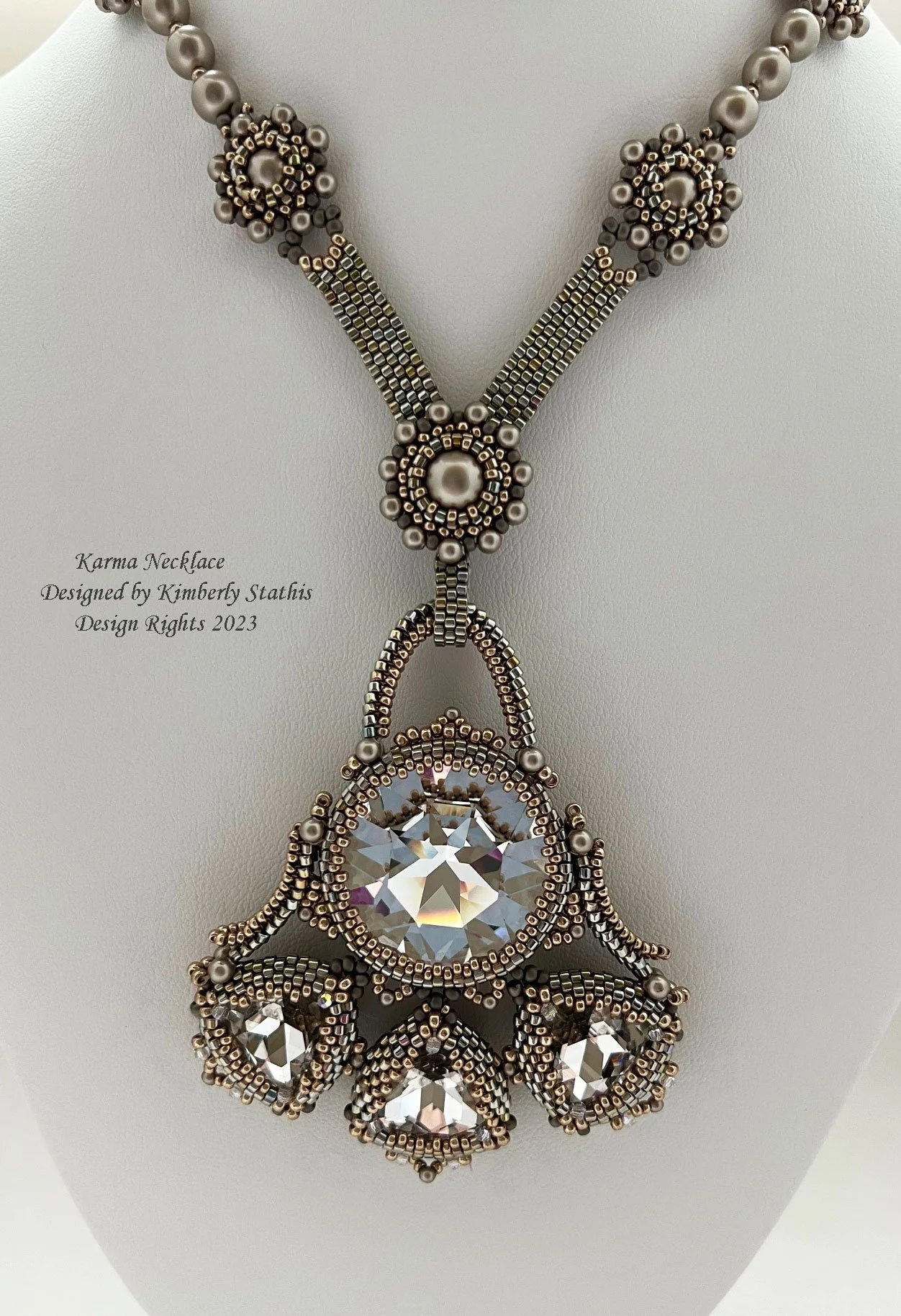 Close-up of a beaded necklace with a large central clear gemstone and silver, pearl, and black beads, labeled as 'Karma Necklace' designed by Kimberly Stathis in 2023.