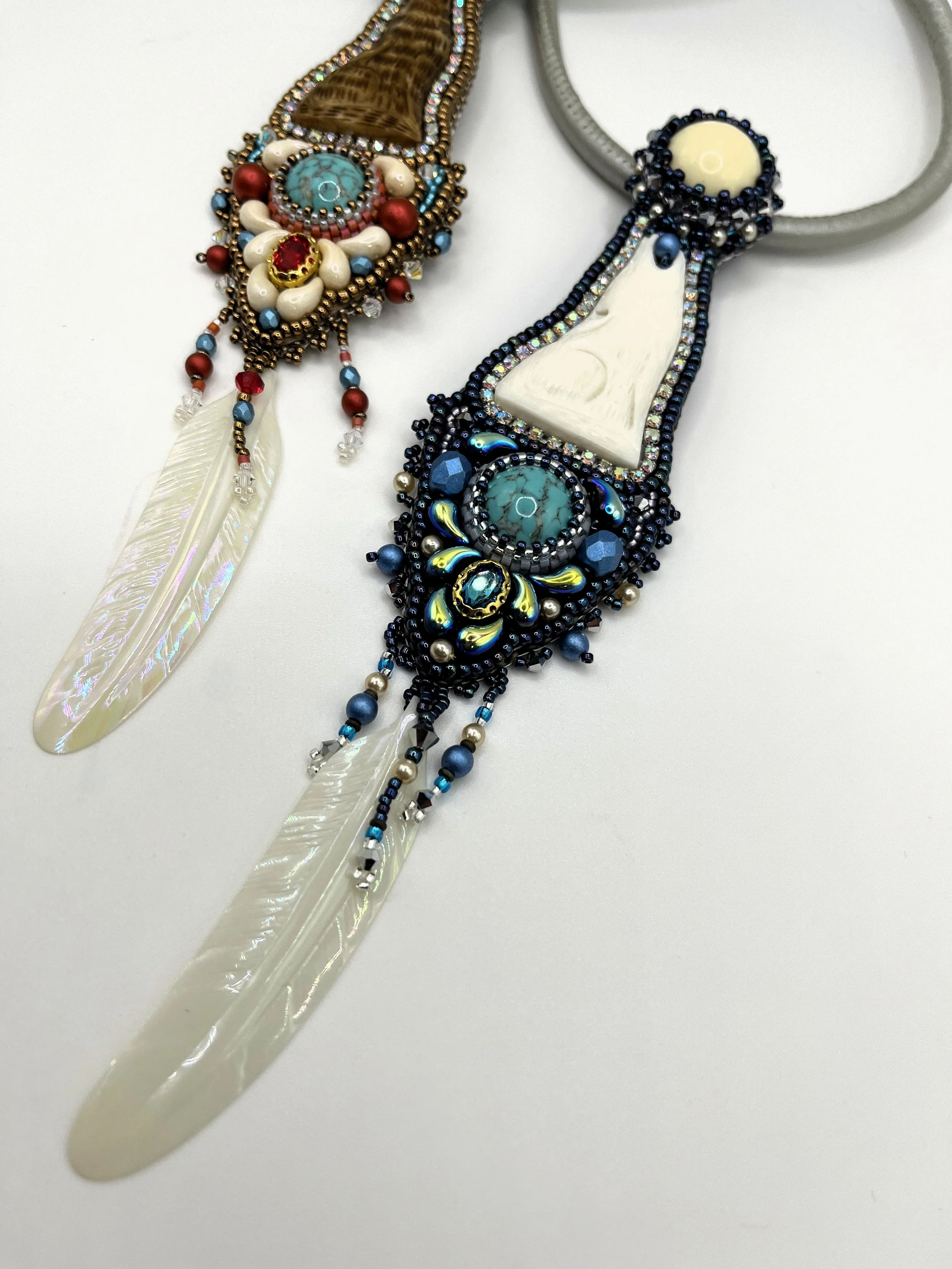 Close-up of two decorative necklaces with beads, gemstones, feathers, and beaded fringe, laid on a white surface.