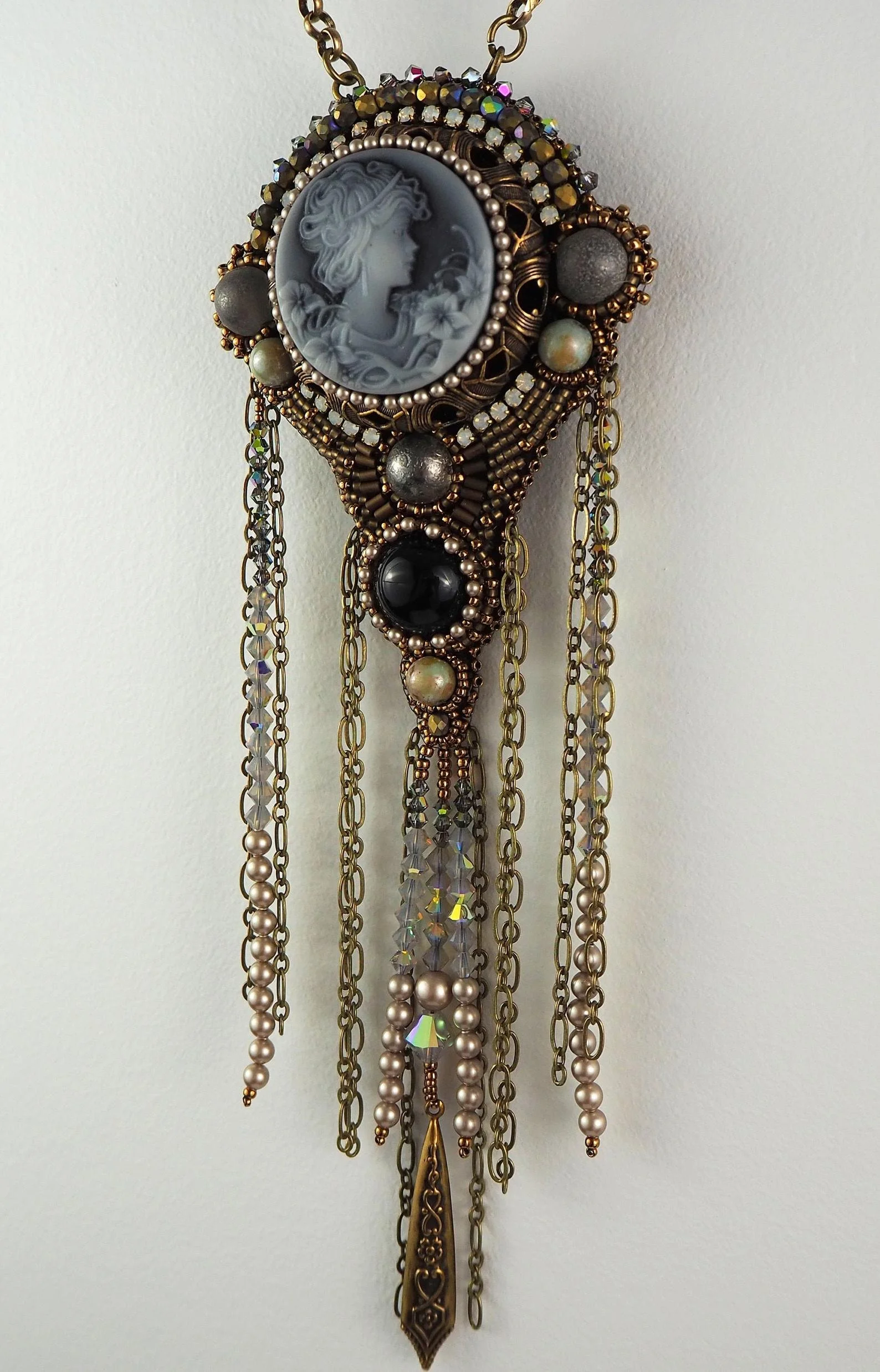 A decorative hanging jewelry piece with a cameo portrait of a woman with flowing hair, surrounded by various chains, beads, and ornamental elements in gold, silver, and pearl tones.