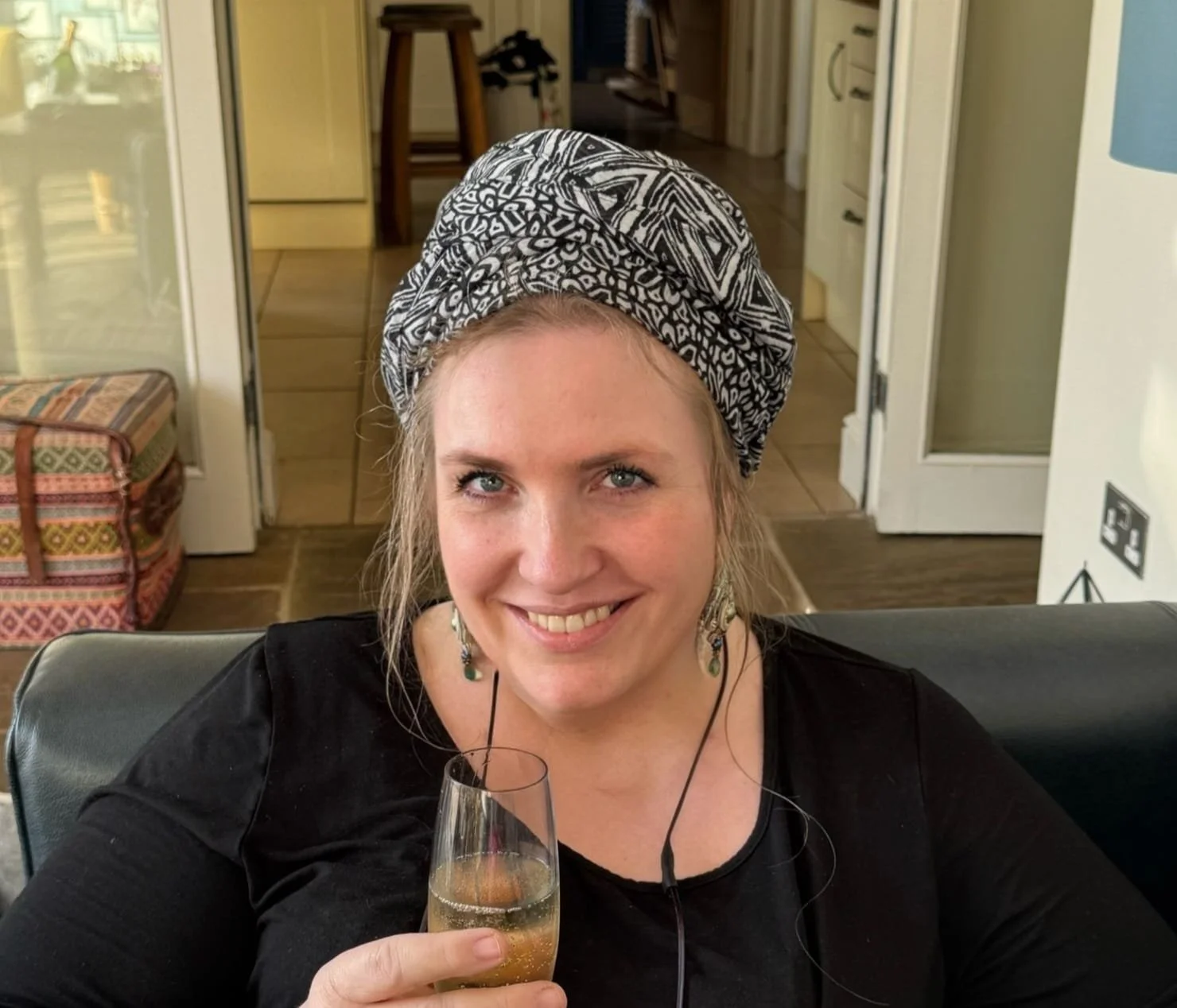 A woman with blonde hair and blue eyes smiling while holding a glass of champagne. She is wearing a black top, earrings, and a patterned headscarf. She is sitting on a black leather sofa in a cozy room with wooden flooring, a patterned suitcase, and a door behind her.