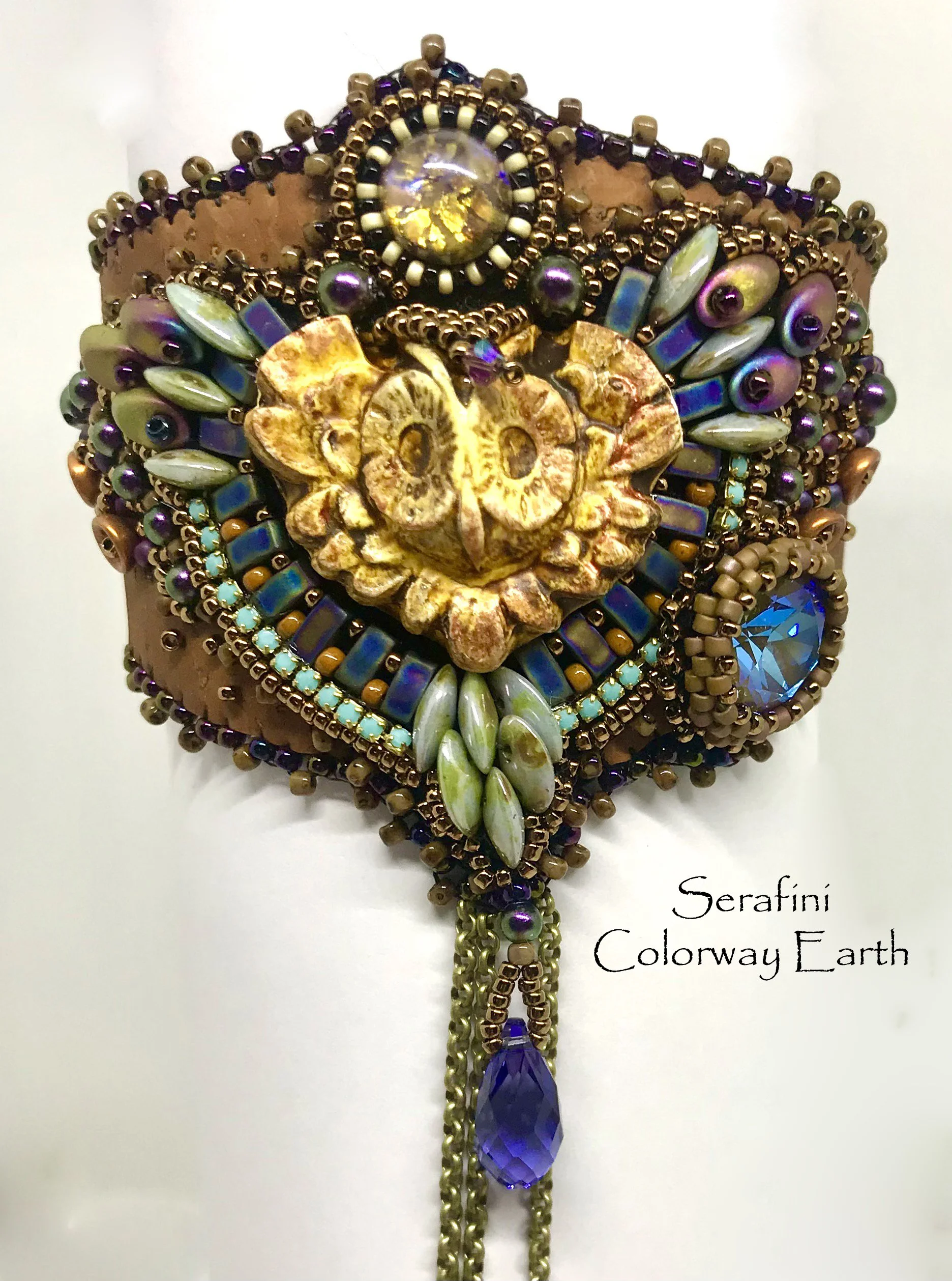 Decorative necklace featuring metallic beads, iridescent stones, and a central ornate lion face with gemstone accents. The necklace has dangling chains with a large purple gemstone at the bottom. The background includes some text.