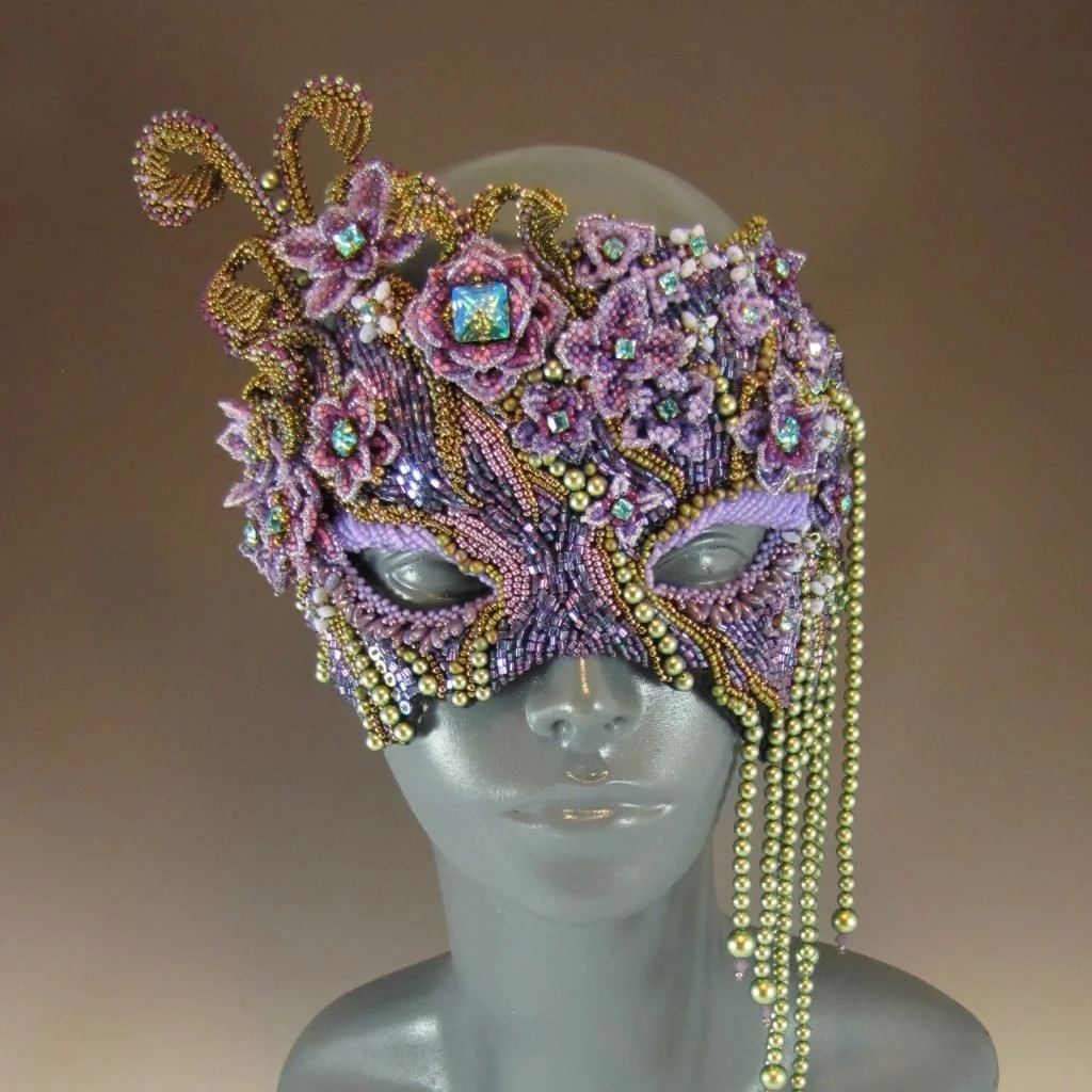 An ornate mask decorated with pink and purple beads, gold accents, and rhinestones, displayed on a mannequin head.