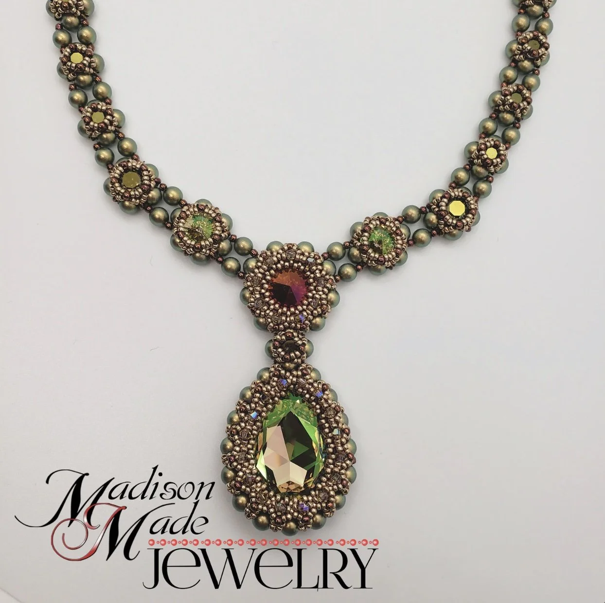 A beaded necklace with multicolored gemstones and intricate beading, featuring a large green faceted pendant, with the text 'Madison Made Jewelry' at the bottom left corner.