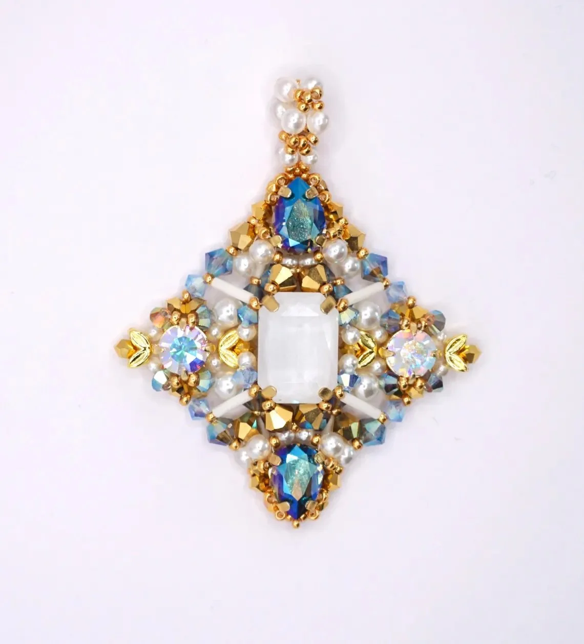 Decorative jewelry piece with pearls, blue and clear crystals, gold accents, and a large rectangular central white gem, arranged symmetrically on a white background.