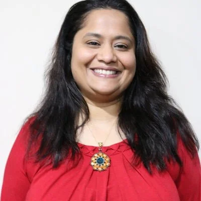 Smiling woman with long dark hair wearing a red top and a colorful pendant necklace against a plain background.