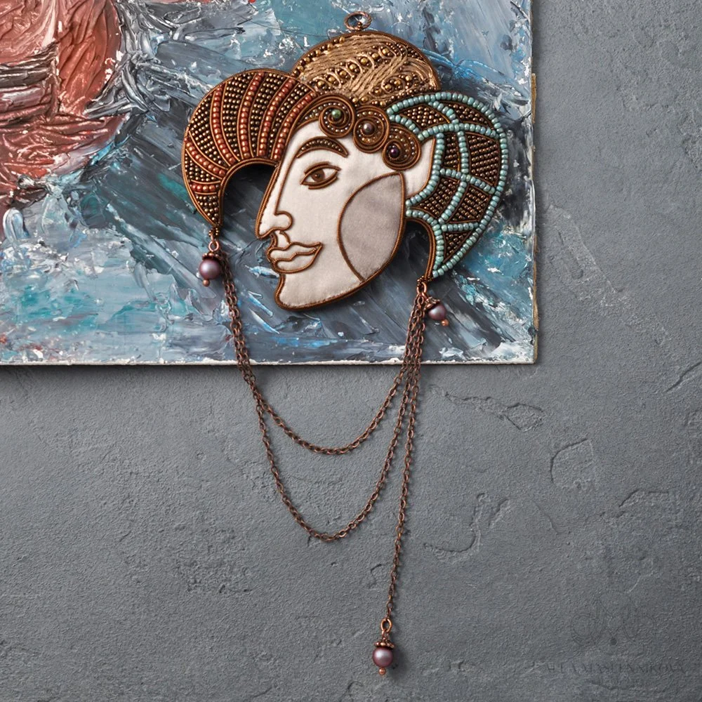 A decorative wall hanging necklace with a face design, featuring intricate beading and chain details, mounted on a textured gray wall with an abstract painting in the background.