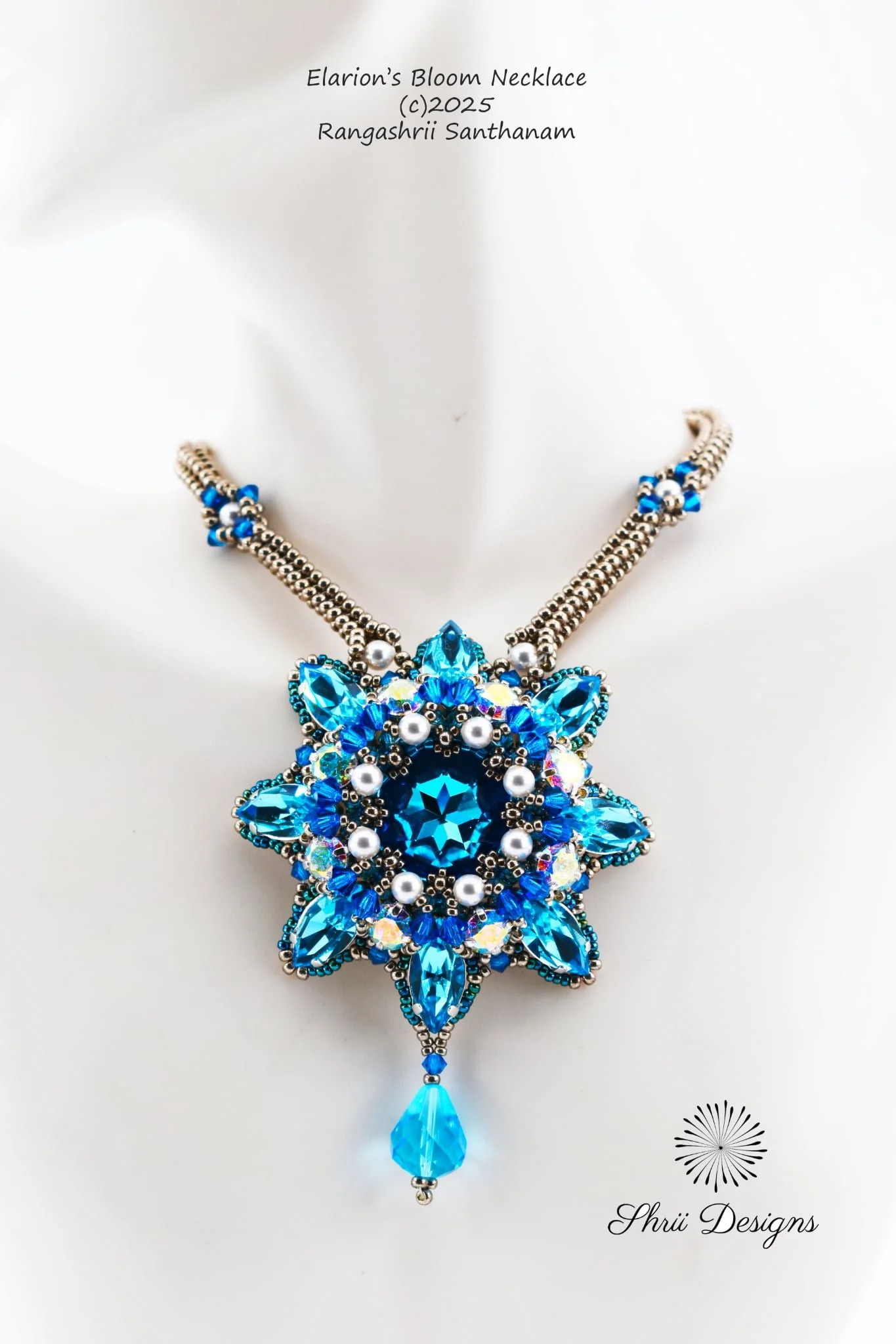 A blue and silver floral necklace with a large blue teardrop pendant, chain links, and small beads on a white display card. The card has text reading "Elarion's Bloom Necklace" and a copyright notice for 2025 by Rangashrii Santhanam, with a logo and 