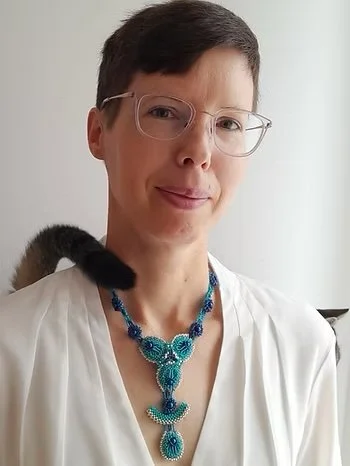 Portrait of a woman with short dark hair and glasses, wearing a white blouse and a blue beaded necklace, with a black and brown cat on her shoulder.