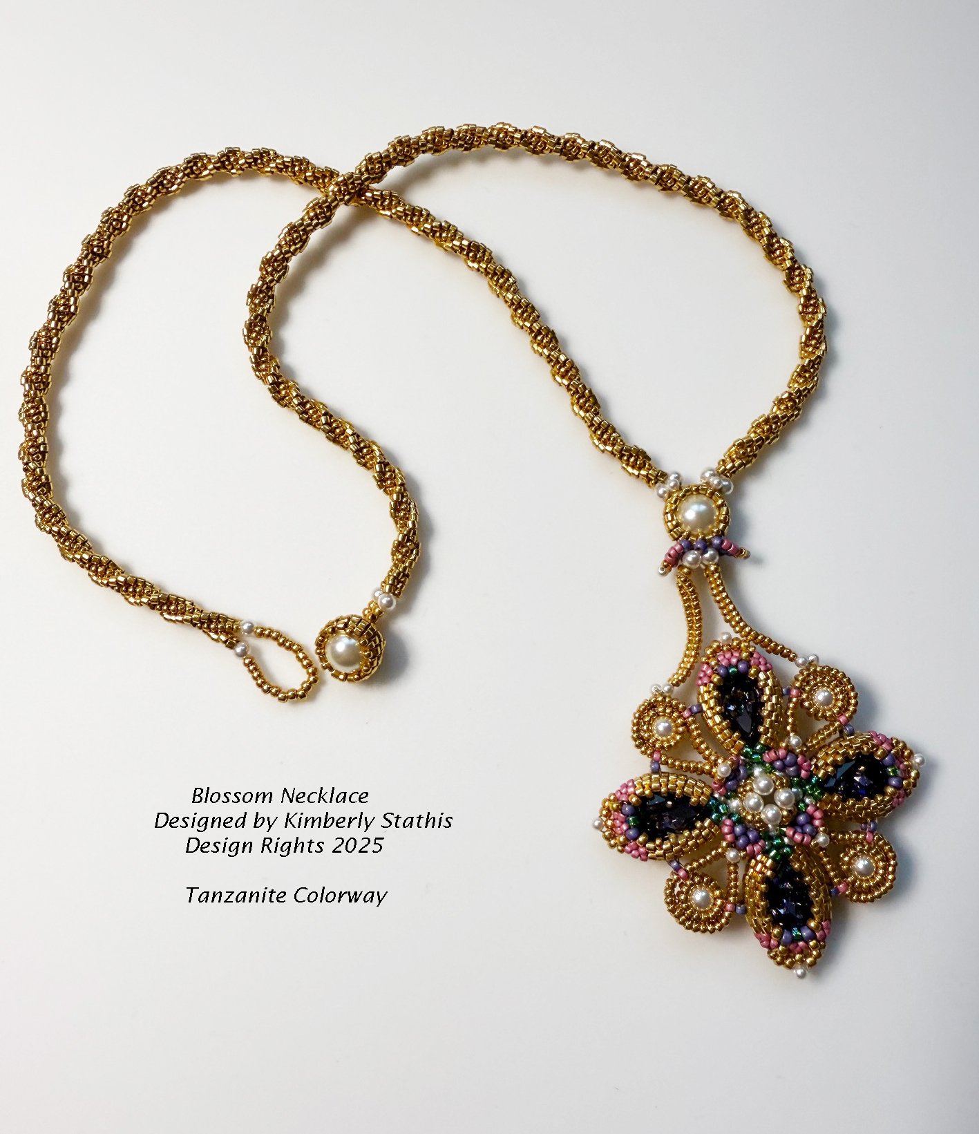A gold beaded necklace called Blossom Necklace with a floral pendant featuring black, pink, green, and white beads, designed by Kimberly Stathis, called Tanzanite Colorway, 2025