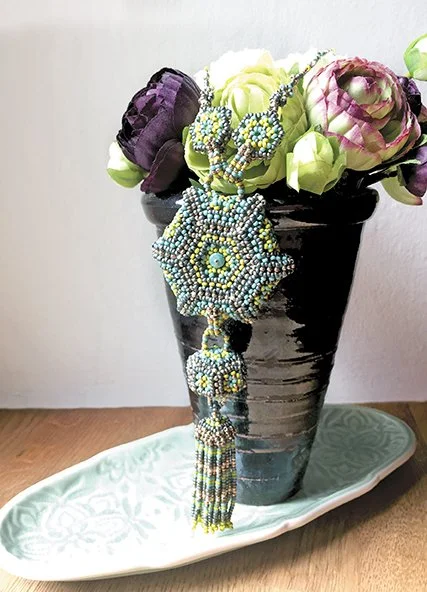Beaded jewelry displayed on a flower arrangement with white, purple, and pink flowers in a black vase on a wooden surface, with a white patterned tray underneath.