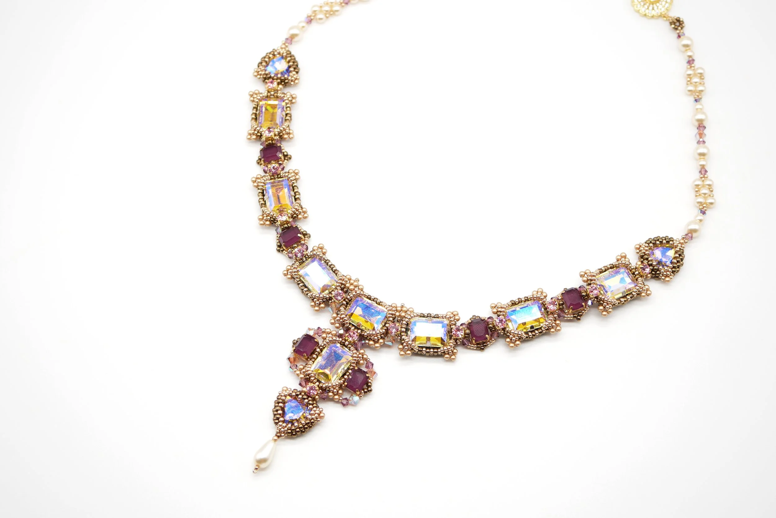 A gold necklace adorned with rectangular and round purple and iridescent stones, featuring pearl accents and a drop pearl pendant.