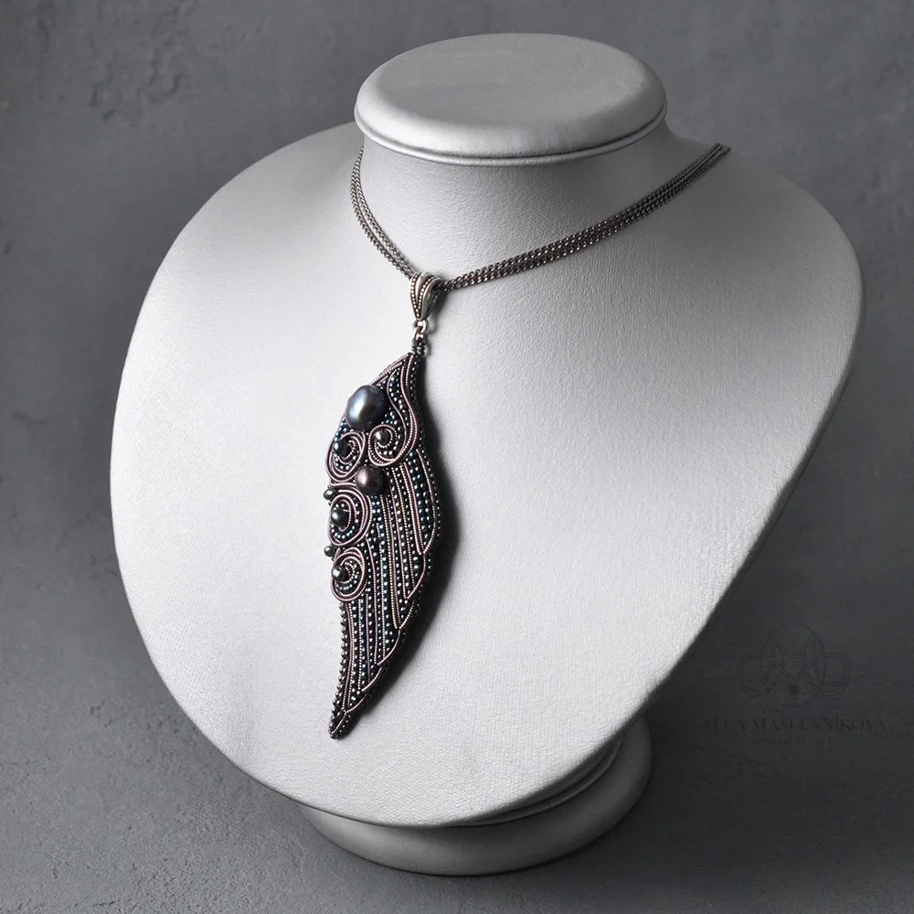 Silver necklace with a large, ornate feather-shaped pendant, adorned with beads and intricate swirl designs, displayed on a white jewelry bust.