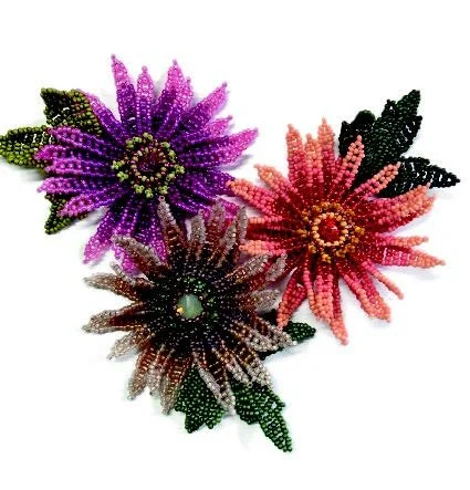 Up close view of three colorful beaded flower brooches in purple, pink, and brown with green beads for leaves on a white background.