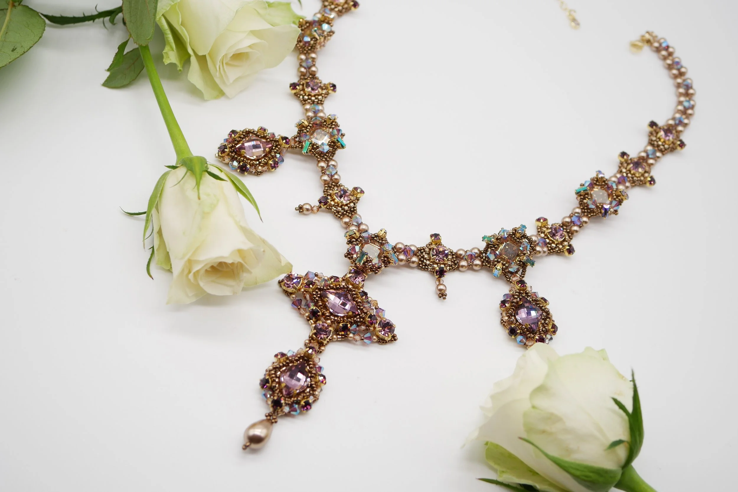 A jewelry set consisting of a necklace and earrings, decorated with pink, purple, and iridescent gemstones, laid on a white surface next to white roses with green leaves.