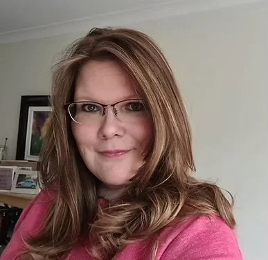 A woman with long, wavy, light brown hair and glasses, wearing a pink top, smiling indoors with framed pictures on the wall behind her.