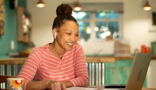A woman with curly hair in a bun, wearing a pink striped shirt, smiling while looking at her laptop and wearing wireless earbuds in a cozy kitchen or cafe setting.