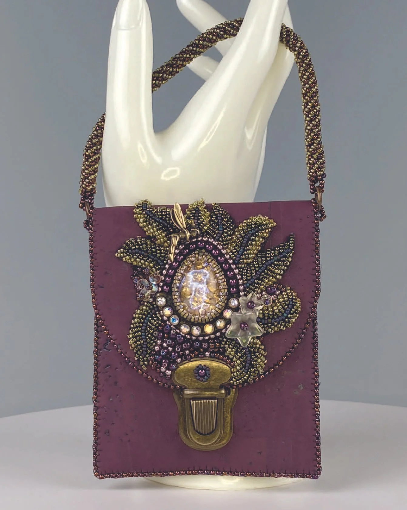 A decorative jewelry box with a beaded necklace draped over a mannequin hand, featuring an elaborately decorated clasp with beads, crystals, and a small charm, all set against a purple fabric background.