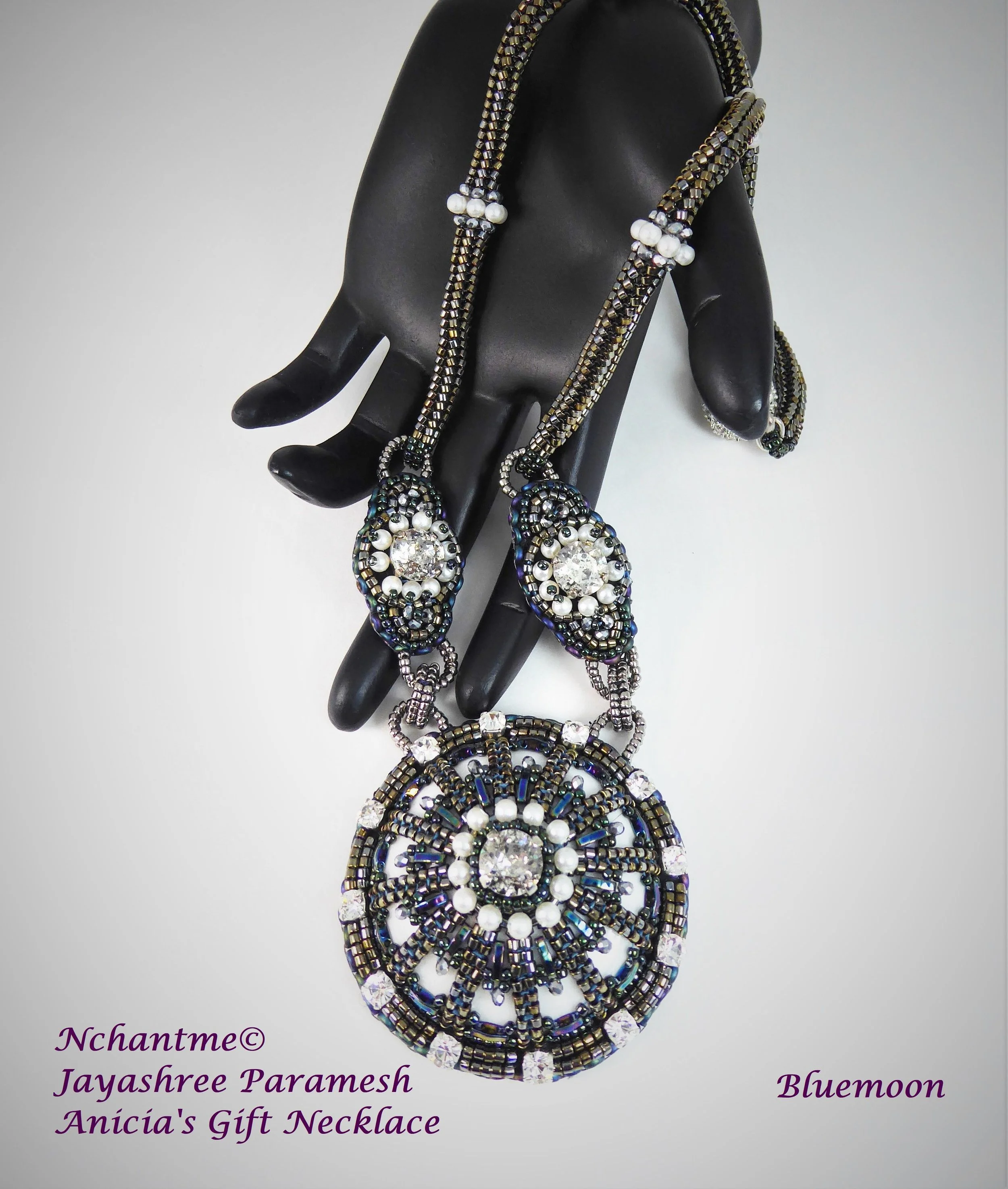 A black hand mannequin displaying a beaded necklace with intricate circular designs, beads in various shades of blue, black, white, and iridescent, adorned with faux pearls. Text in purple at the bottom left corner reads: 'Nchantme© Jayashree Parames