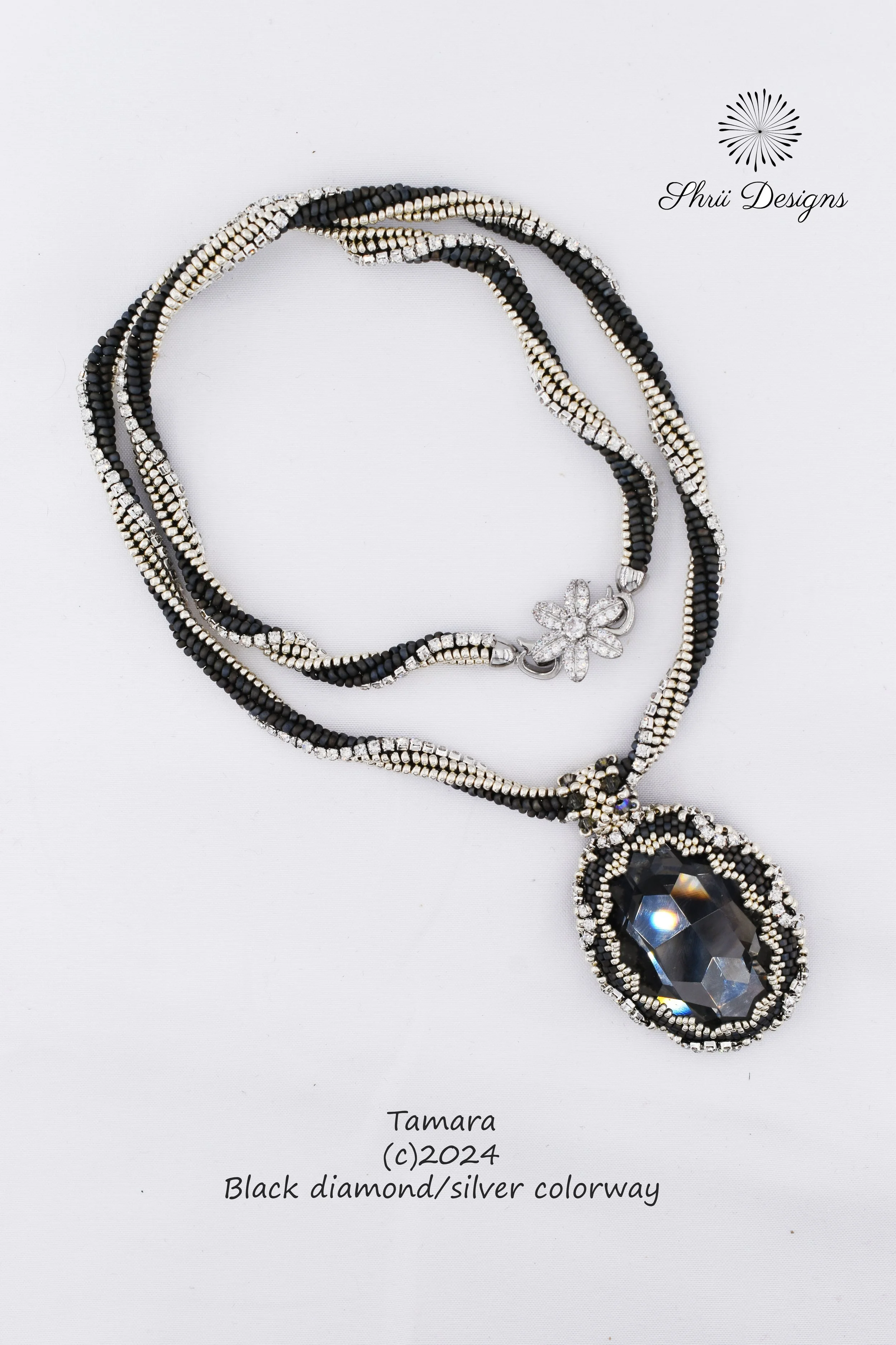 A beaded necklace with black beads and silver beads, featuring a large black diamond-like pendant with a silver flower design. The necklace is labeled as designed by Tamara in 2024, with a description of black diamond and silver colorway. The backgro